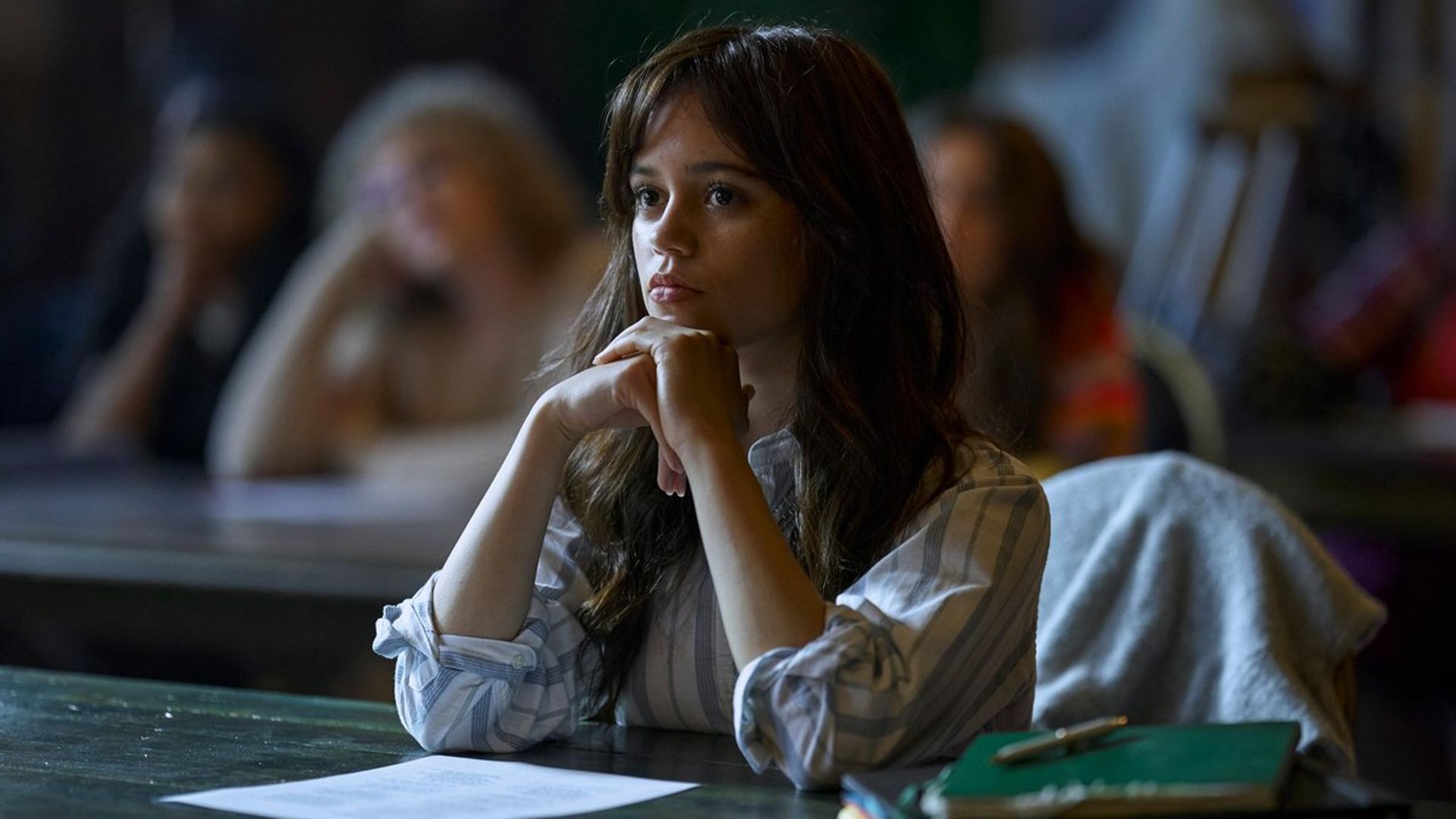 Jenna Ortega w białym swetrze siedzi przy ławce szkolnej ze skupionym wyrazem twarzy