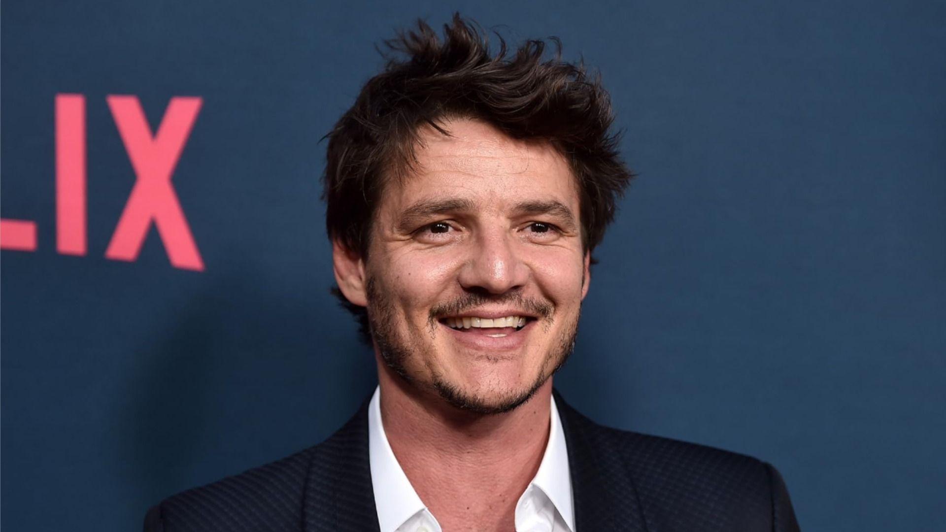 Aktor Pedro Pascal w czarnej kurtce uśmiechający się na niebieskim tle