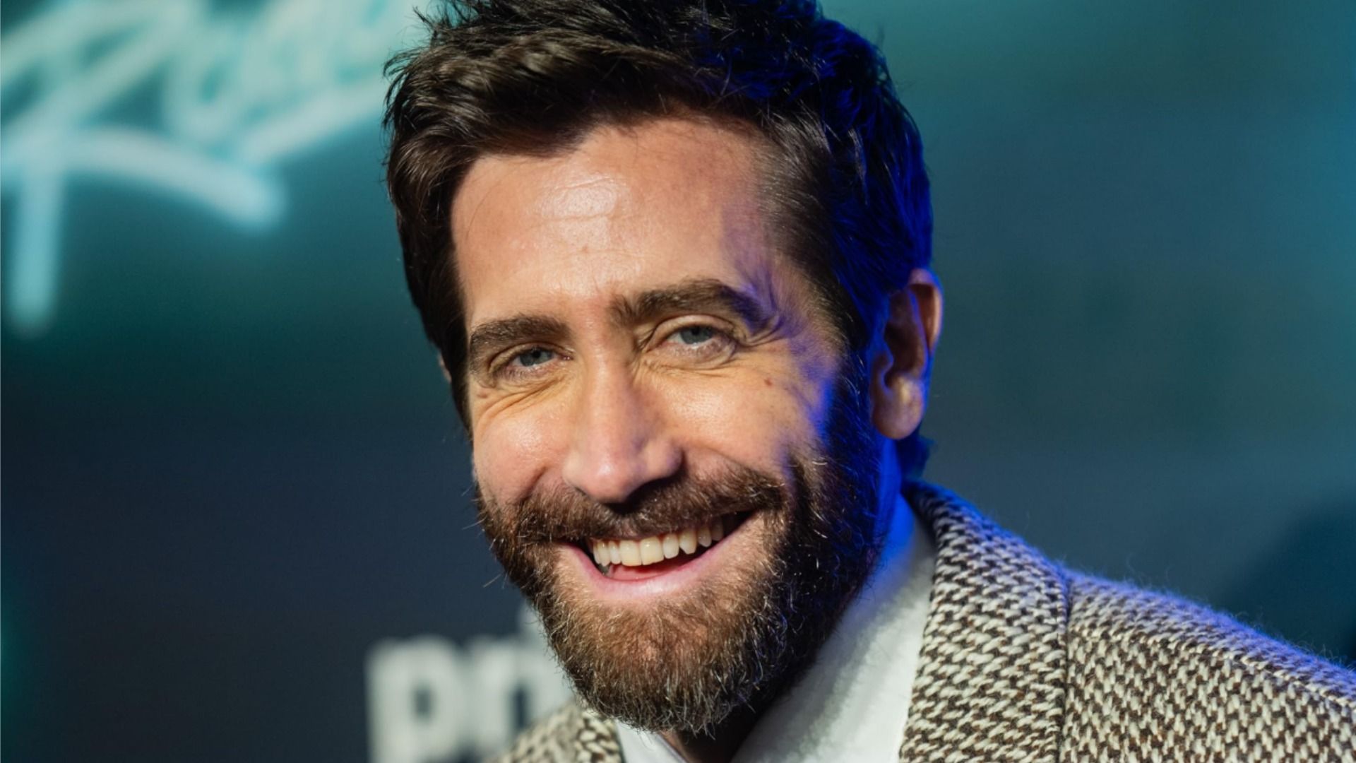 Aktor Jake Gyllenhaal w szarej marynarce uśmiecha się na niebieskim tle