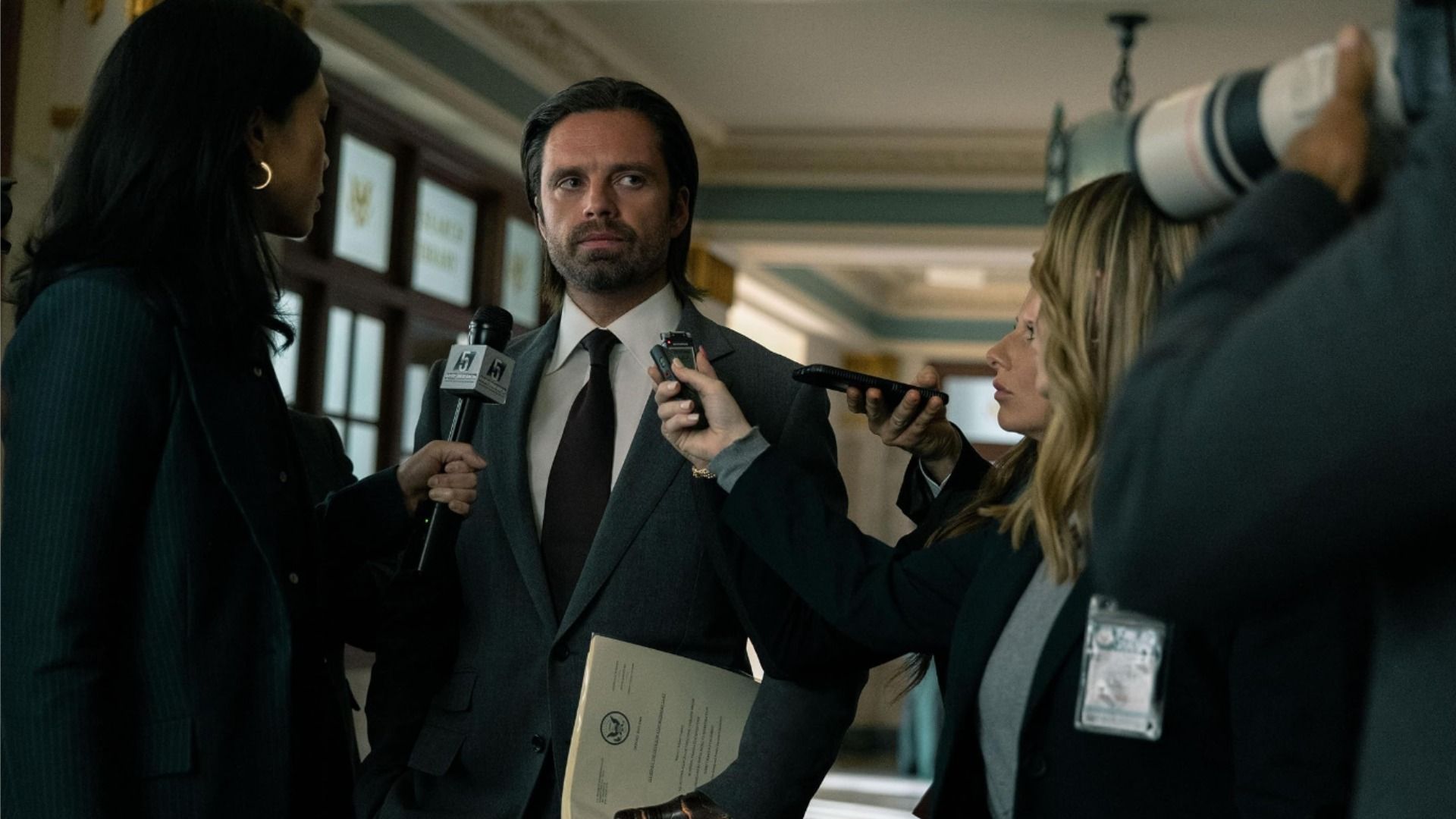 Aktor Sebastian Stan w czarnym garniturze z długimi włosami w otoczeniu dziennikarzy