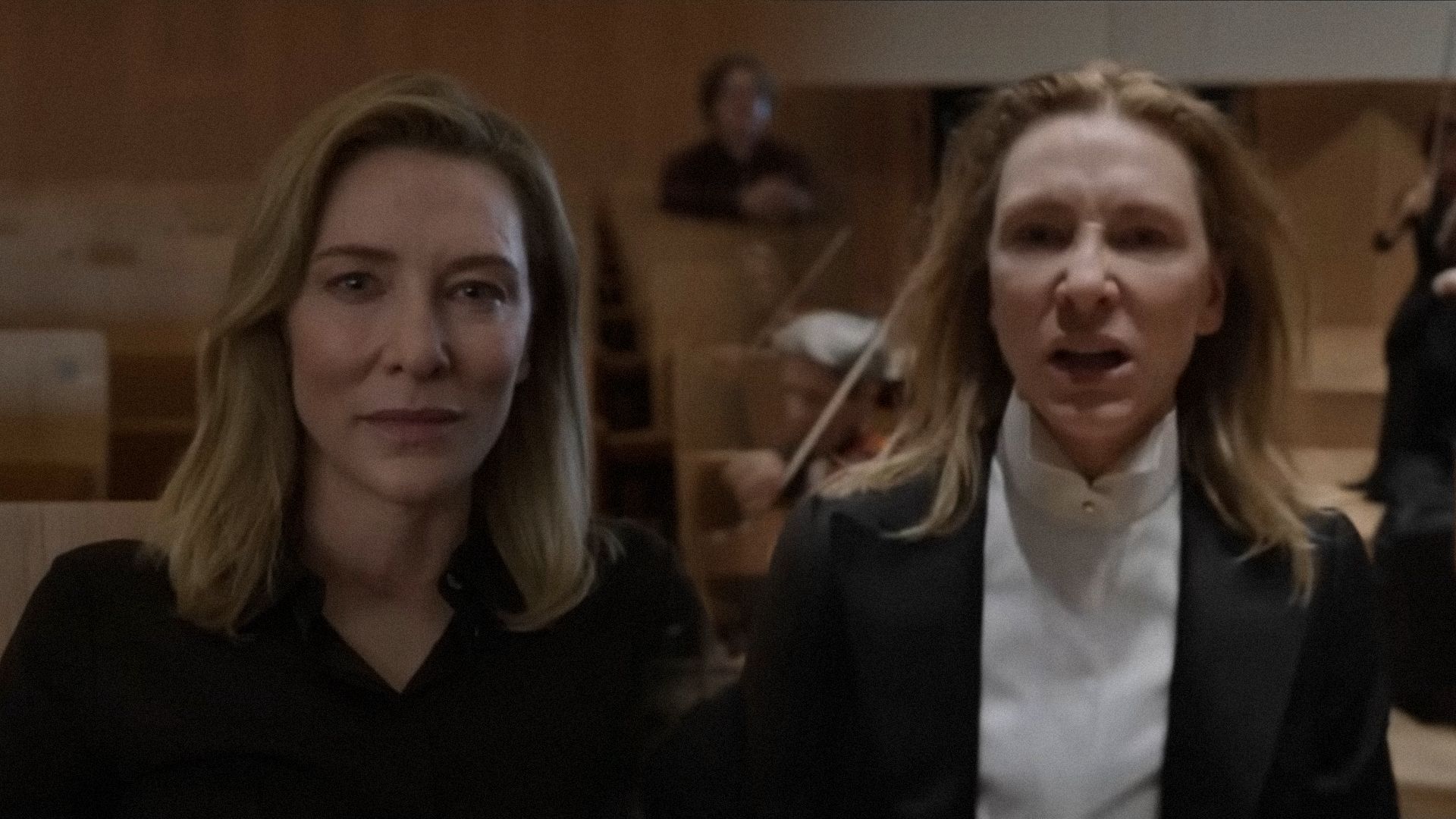 Aktorka Cate Blanchett w czarnym garniturze patrzy groźnie w kamerę na tle skrzypków