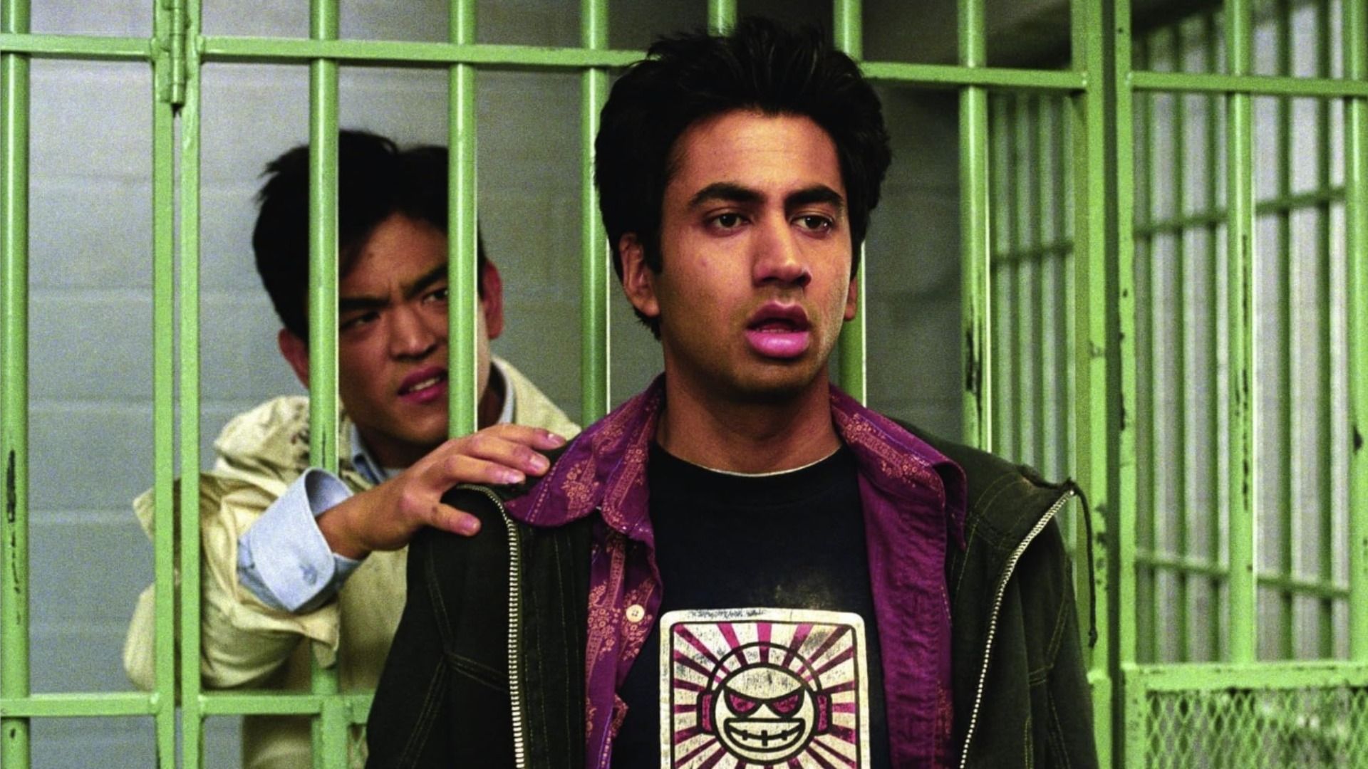 Aktor John Cho stoi za zielonymi kratami więziennymi, aktor Kal Penn stoi na zewnątrz i patrzy w bok
