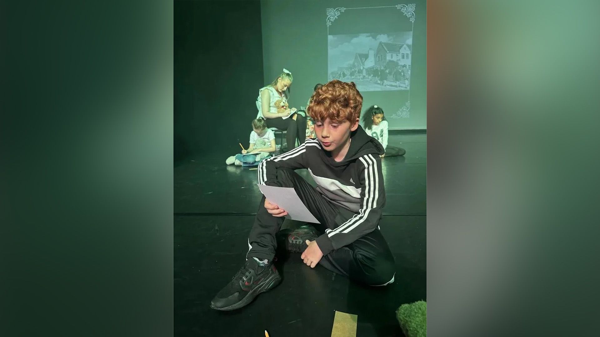 Alastair Stout w dresie siedzący na scenie teatralnej i czytający z kartki