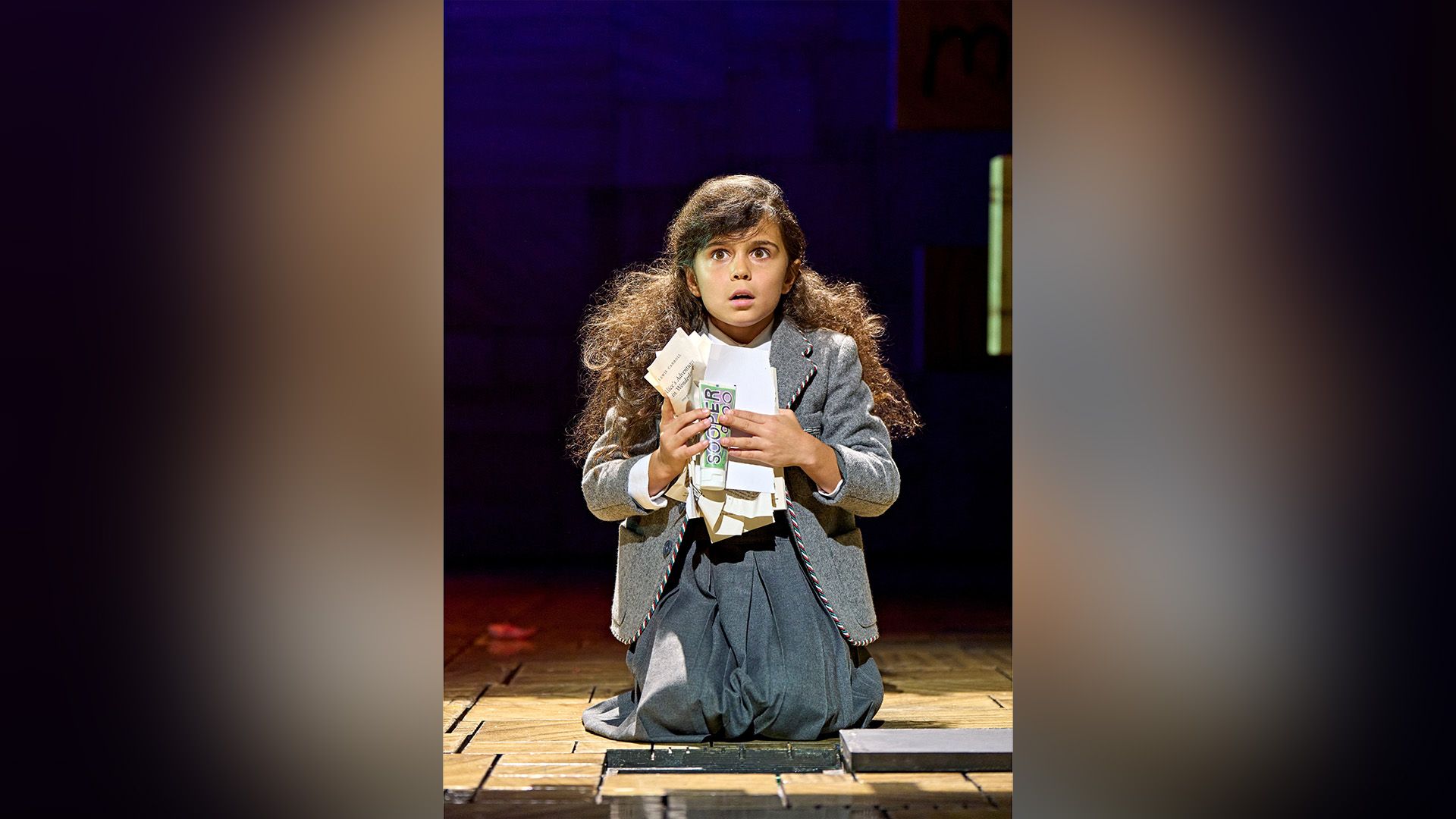 Arabella Stanton w szarej sukience klęczy na scenie teatralnej, trzymając w rękach pogięte kartki papieru