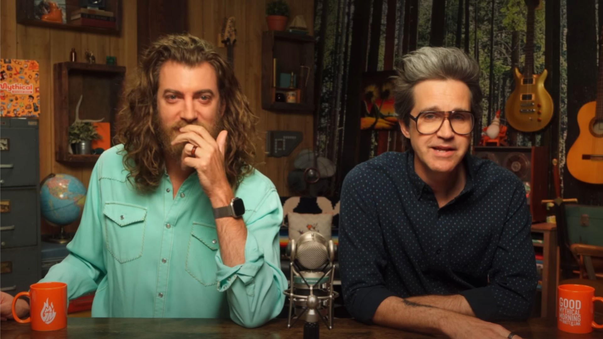 Blogerzy Rhett i Link w koszulach siedzący przy stole z mikrofonem