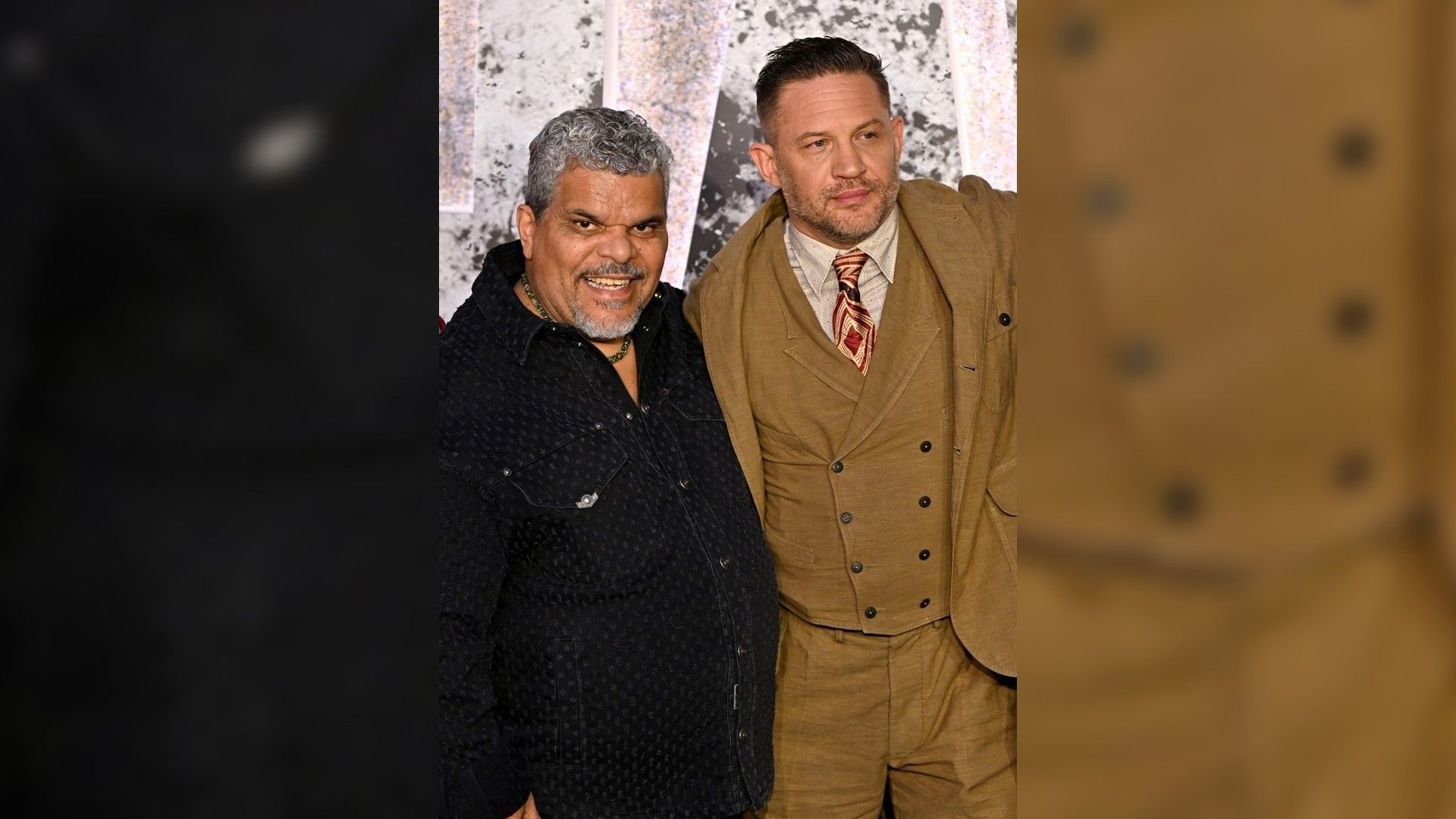 Luis Guzmán i Tom Hardy pozują na czerwonym dywanie: jeden w ciemnej koszuli, drugi w brązowym garniturze z krawatem