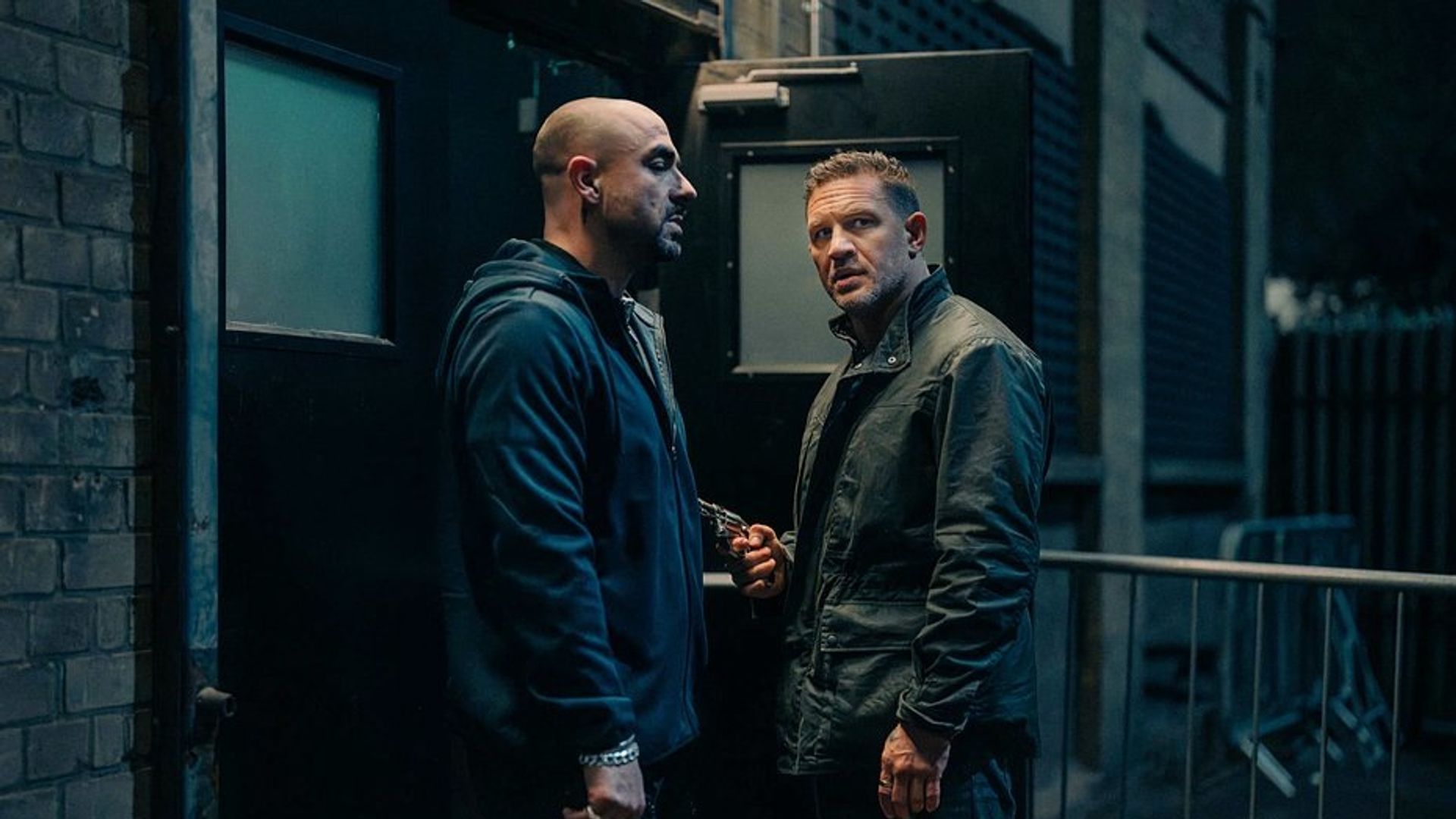 Tom Hardy i inny mężczyzna w ciemnym ubraniu rozmawiają przy czarnym wyjściu nocą, jeden z nich trzyma broń