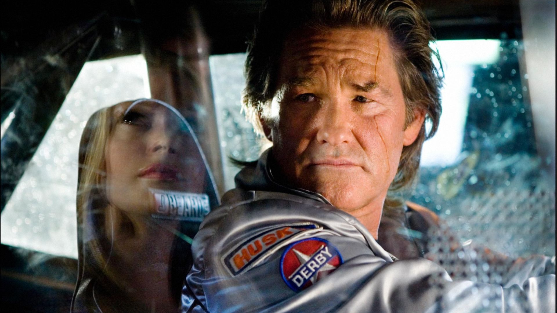 Aktor Kurt Russell w srebrnej kurtce siedzi za kierownicą samochodu i patrzy w bok