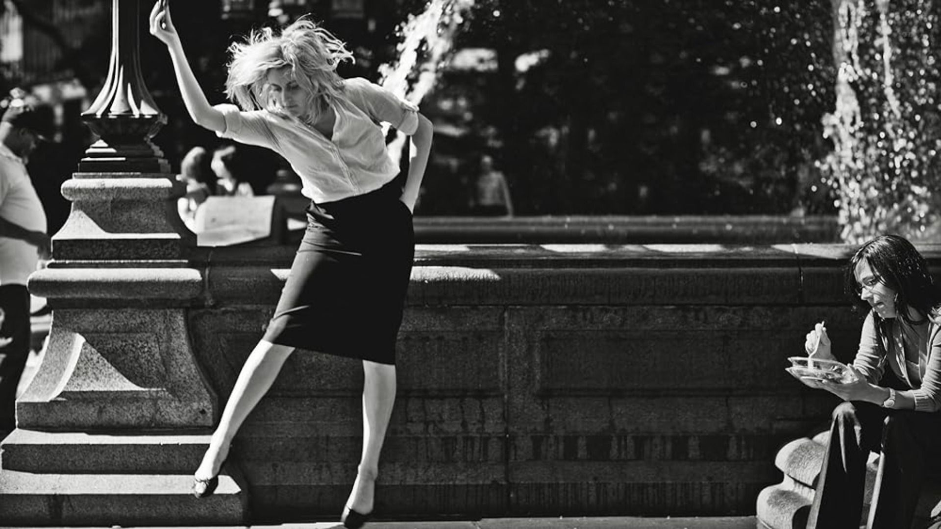 Aktorka Greta Gerwig tańczy w białej bluzce i czarnej spódnicy na tle fontanny