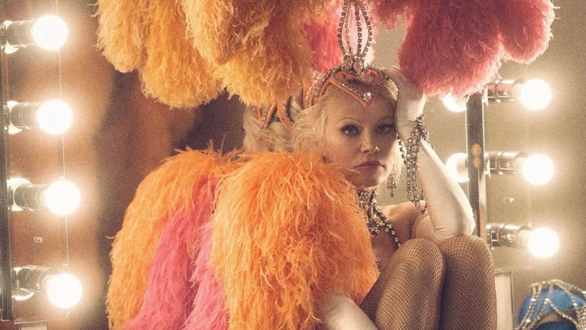 Pamela Anderson w błyszczącym kostiumie scenicznym z piórami i ozdobami siedzi przy lustrze w garderobie, zamyślona, opierając głowę na dłoni