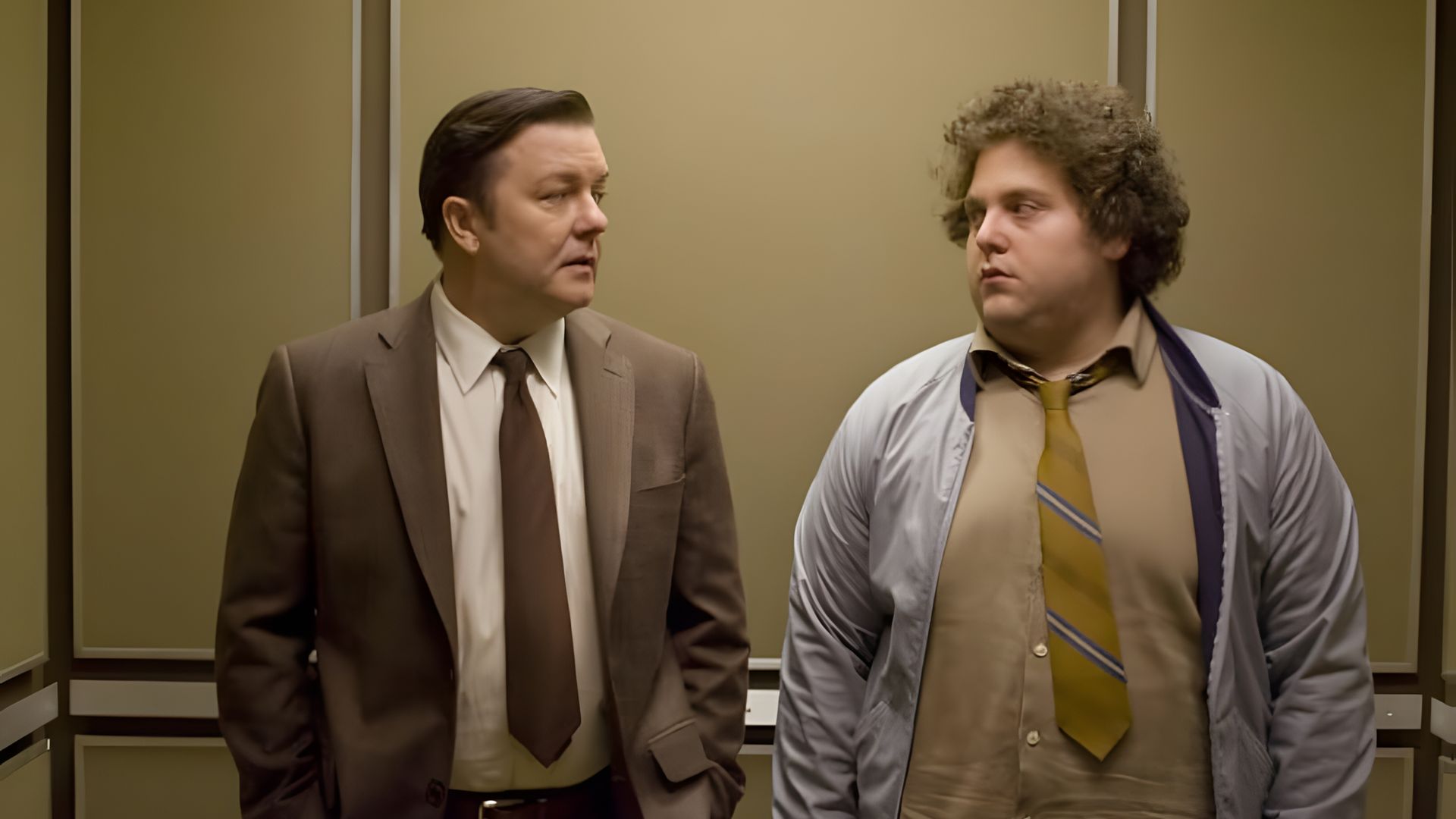 Ricky Gervais i Jonah Hill w garniturach stoją w windzie, patrząc na siebie z poważnymi minami