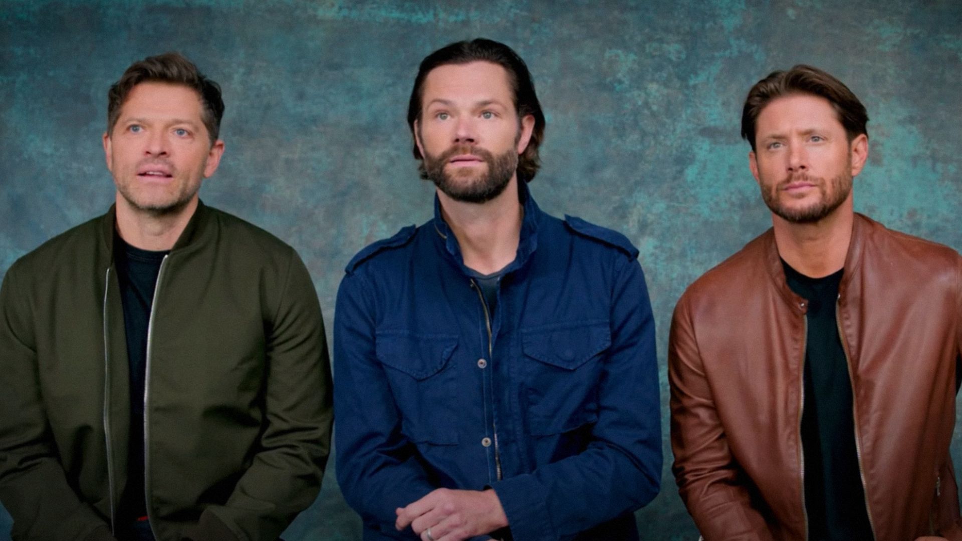 Aktorzy Misha Collins, Jared Padalecki i Jensen Ackles siedzą na tle niebieskiego tła w kolorowych kurtkach