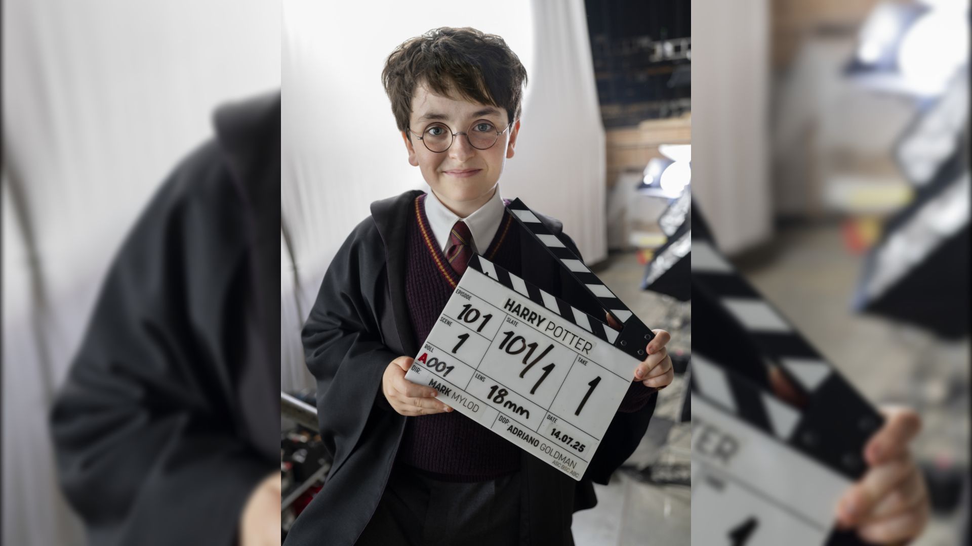 Aktor Dominic McLaughlin w czarnej pelerynie i okularach trzyma klaps filmowy