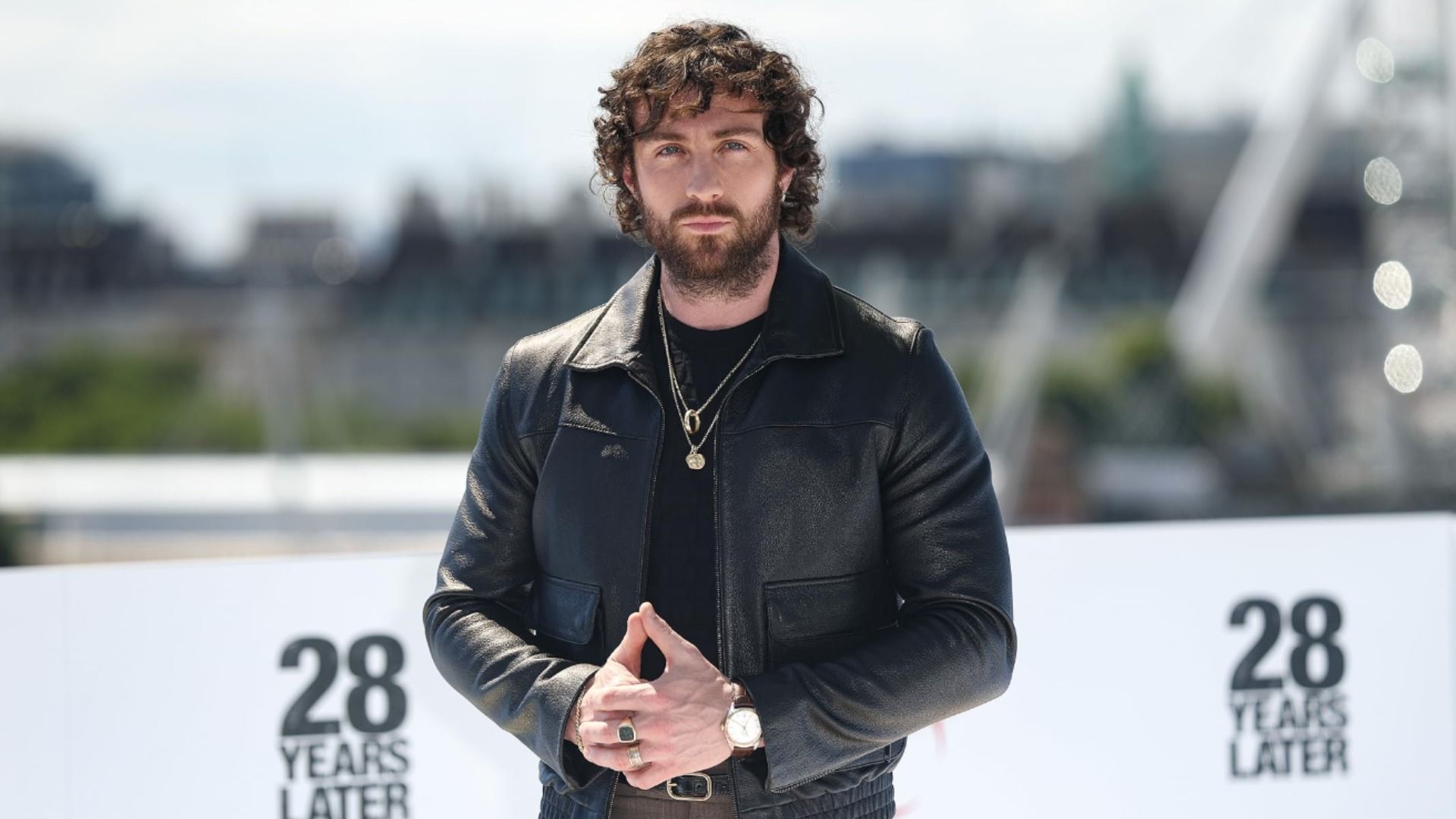 Aktor Aaron Taylor-Johnson w czarnej skórzanej kurtce stoi na tle rozmytego miejskiego tła