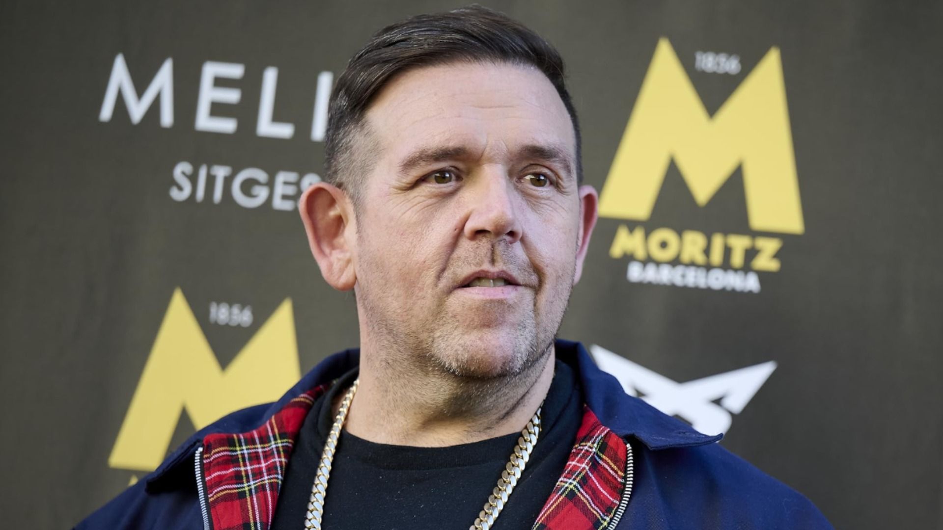 Aktor Nick Frost w niebieskiej kurtce ze złotym łańcuchem na szyi uśmiecha się na tle wystawy
