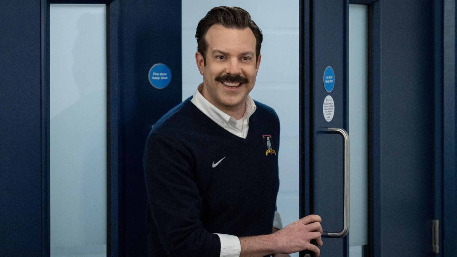 Aktor Jason Sudeikis w niebieskim swetrze i z wąsami stoi przy drzwiach i się uśmiecha