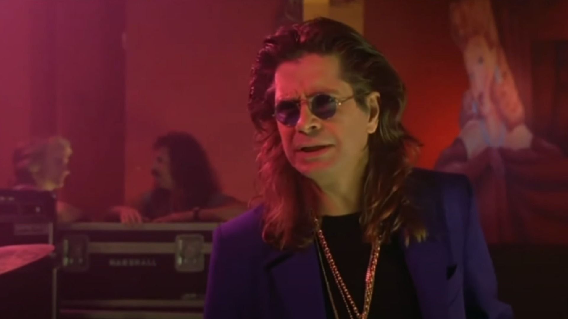 Wokalista Ozzy Osbourne w niebieskiej marynarce i złotym łańcuchu stoi na scenie