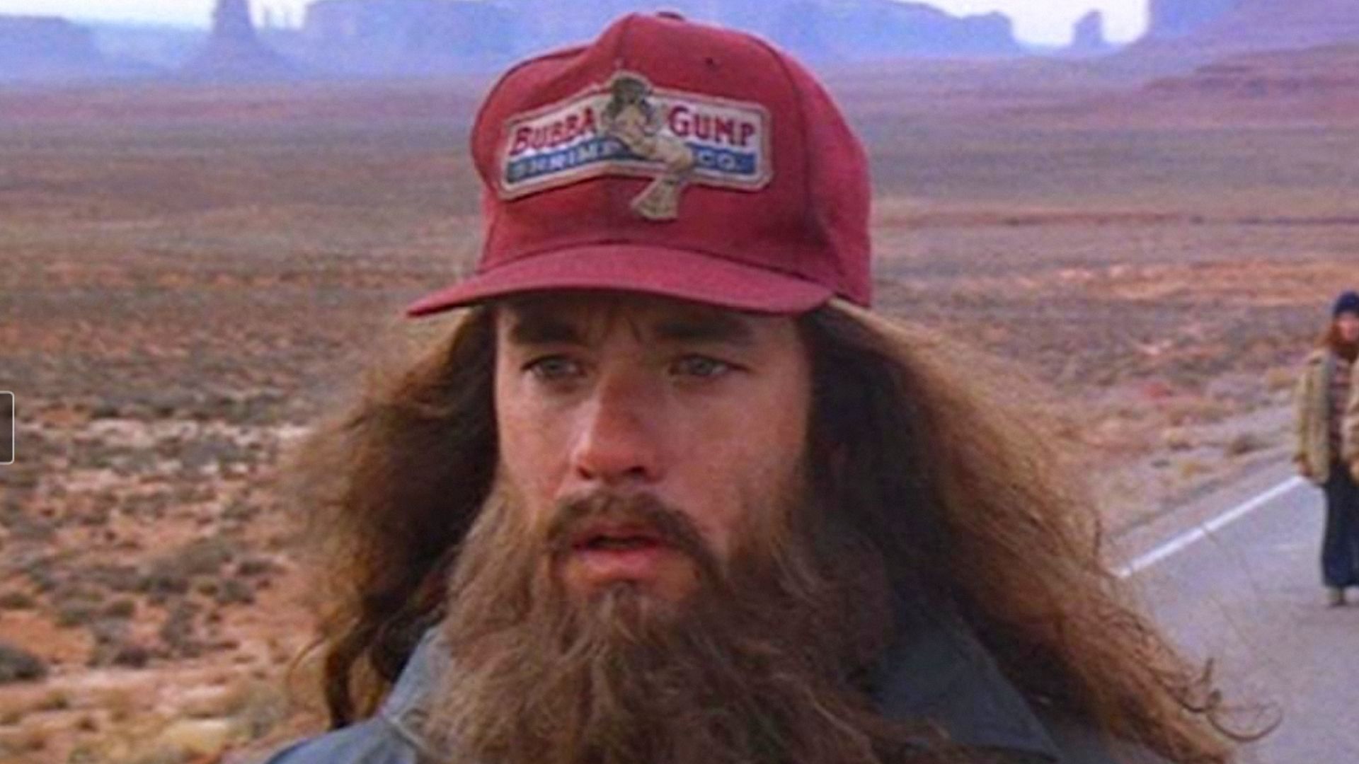 Aktor Tom Hanks z długimi włosami i brodą stoi w czerwonej czapce