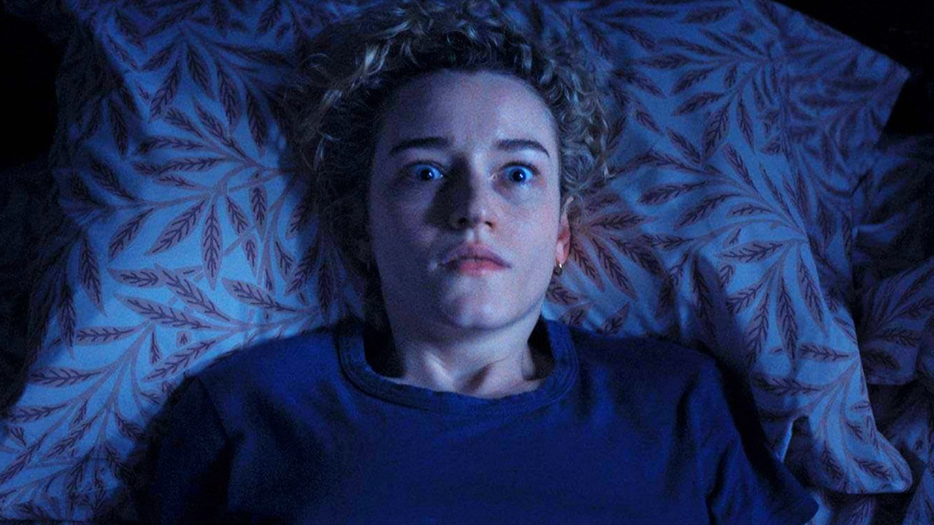 Aktorka Julia Garner w niebieskim swetrze leży na łóżku, przestraszona czymś