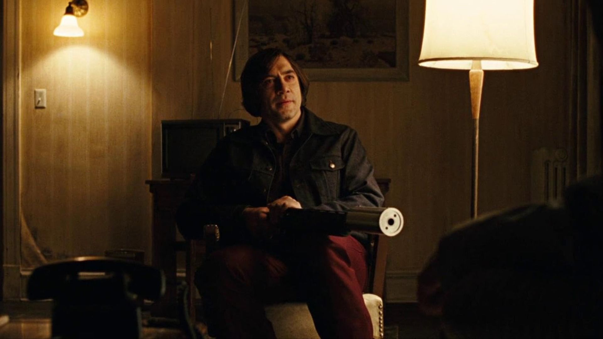 Aktor Javier Bardem z długimi włosami siedzi na krześle, trzymając broń