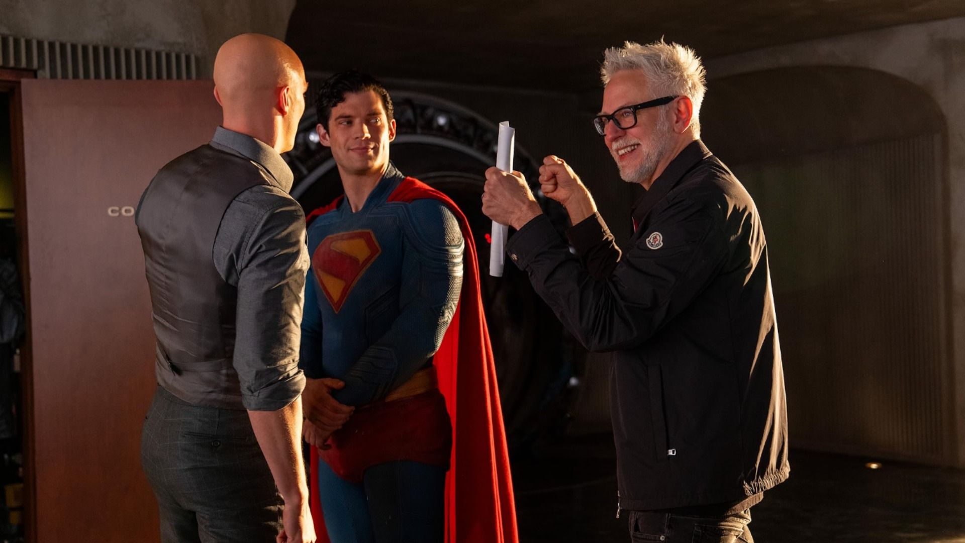 Reżyser James Gunn trzymający papiery rozmawia z aktorem Davidem Corenswet w kostiumie Supermana i z łysym Nicholasem Houltem
