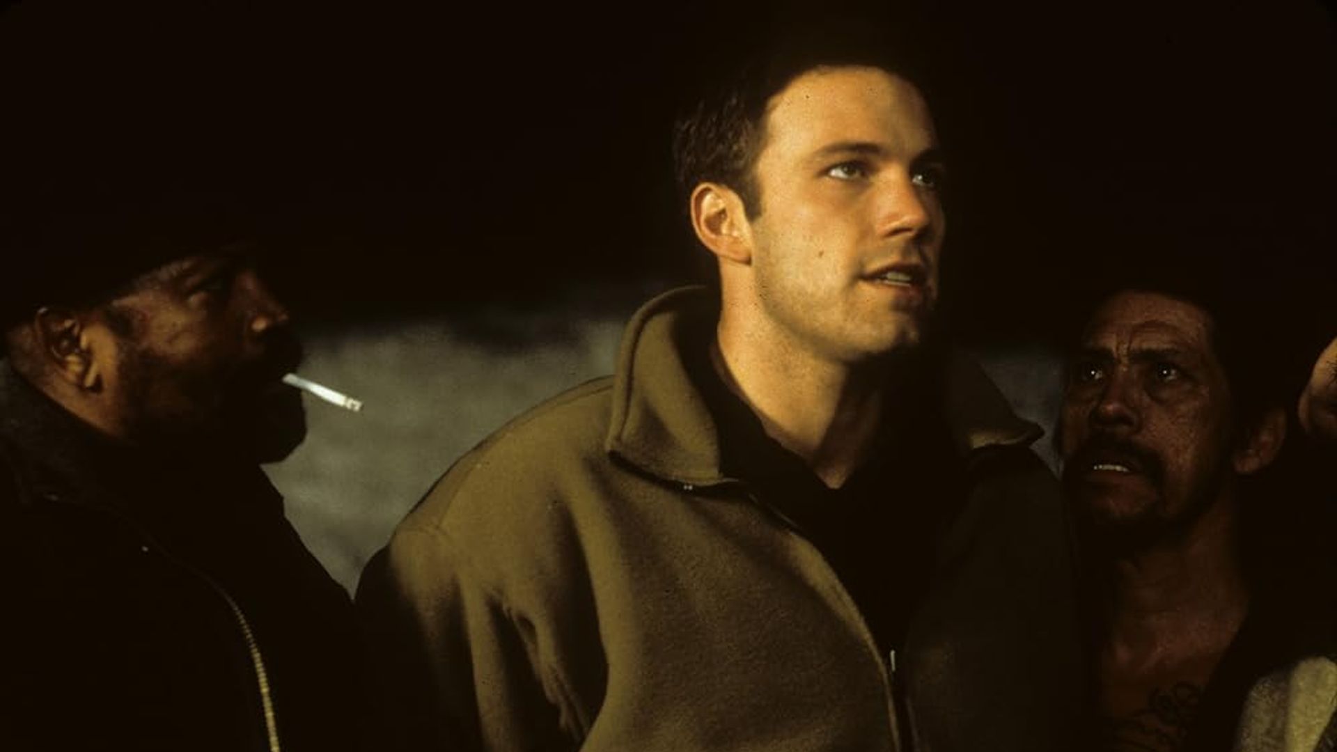 Ben Affleck w beżowej kurtce stoi między dwiema osobami, z których jedna pali, w słabo oświetlonym otoczeniu