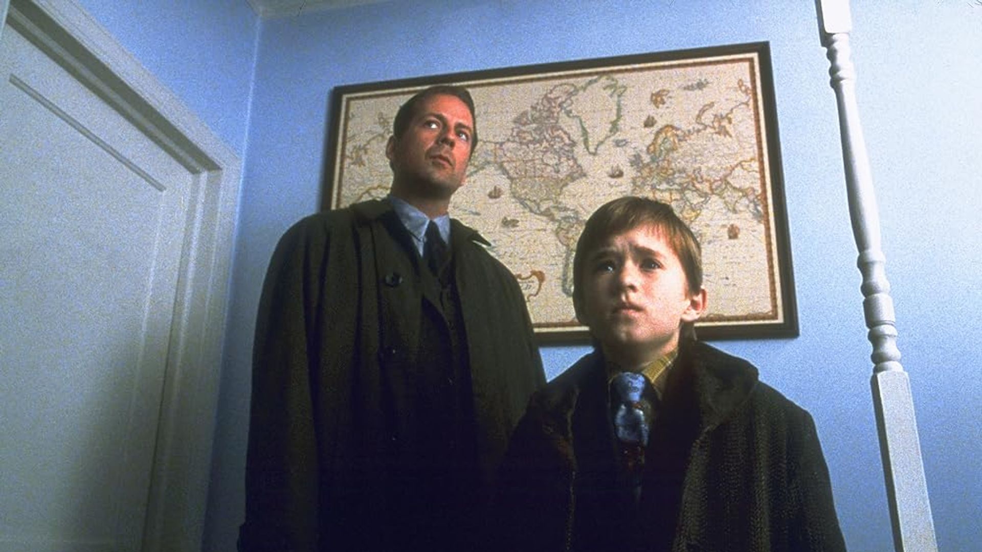 Bruce Willis i Haley Joel Osment stoją w korytarzu z mapą świata wiszącą na ścianie za nimi