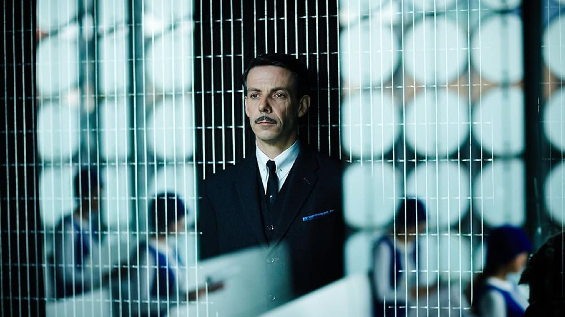 Ethan Hawke w garniturze z wąsami stoi za metalową przegrodą, w tle umundurowani ludzie pracują przy biurkach
