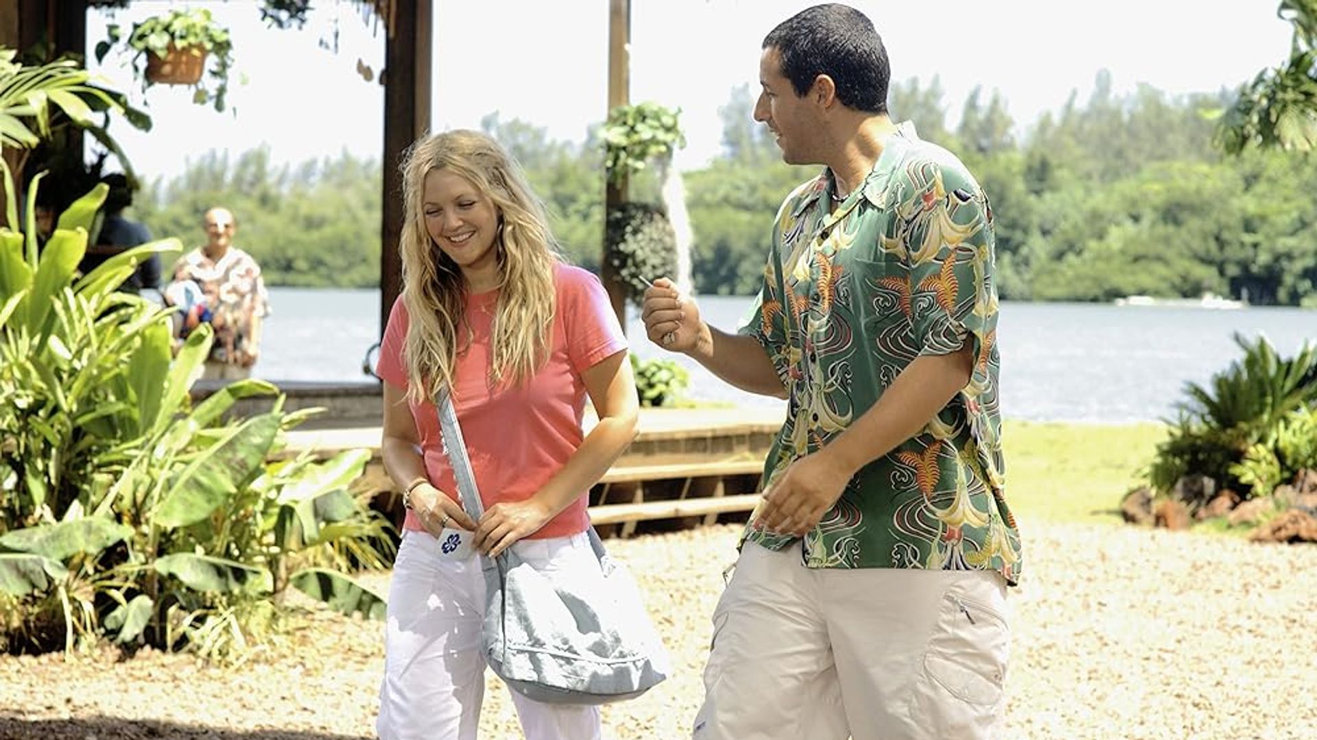 Drew Barrymore i Adam Sandler rozmawiający na słonecznej plaży z zielenią i wodą w tle