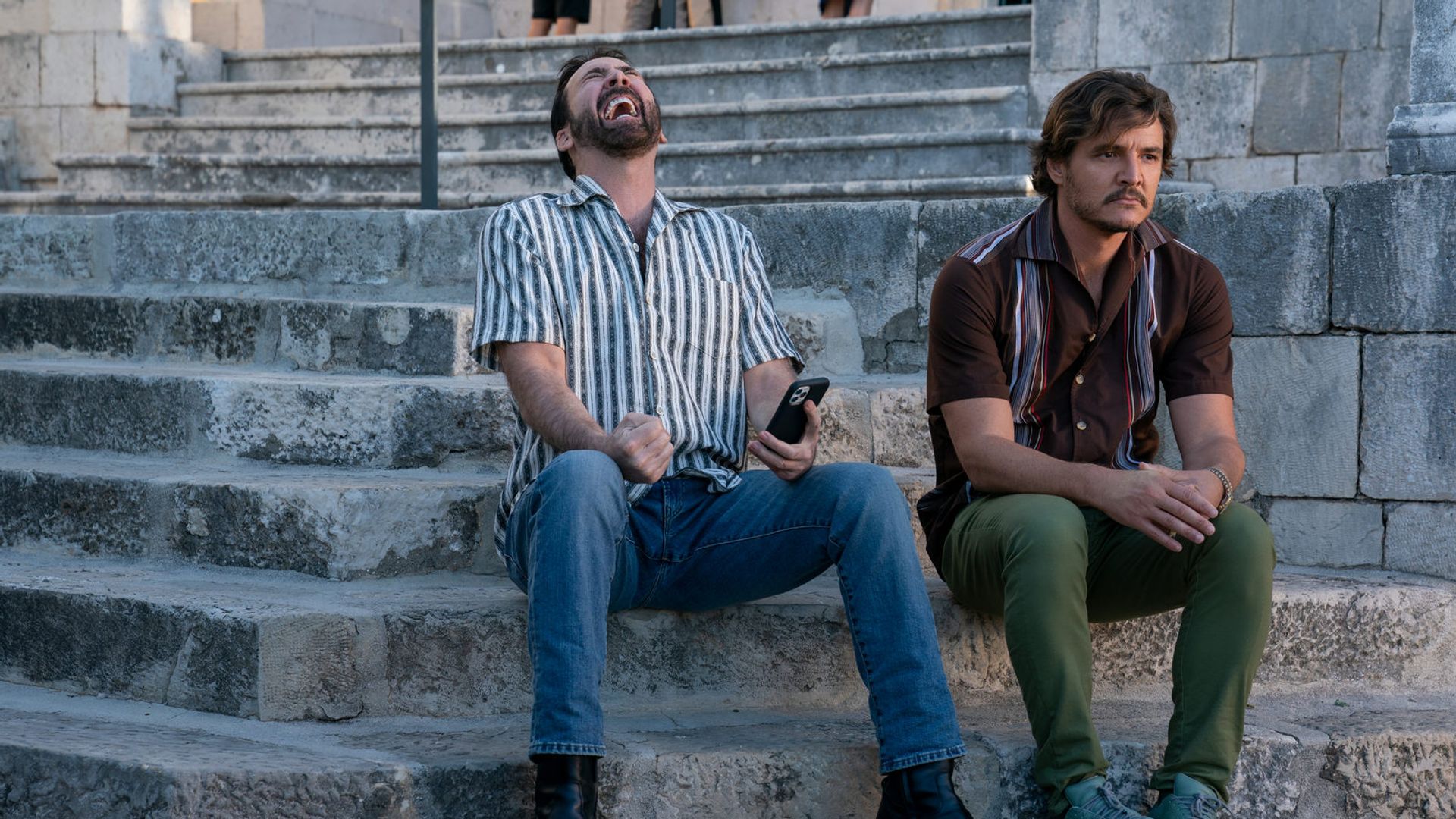Nicolas Cage i Pedro Pascal siedzą na kamiennych schodach: jeden śmieje się z telefonem w ręku, drugi patrzy zamyślony w bok