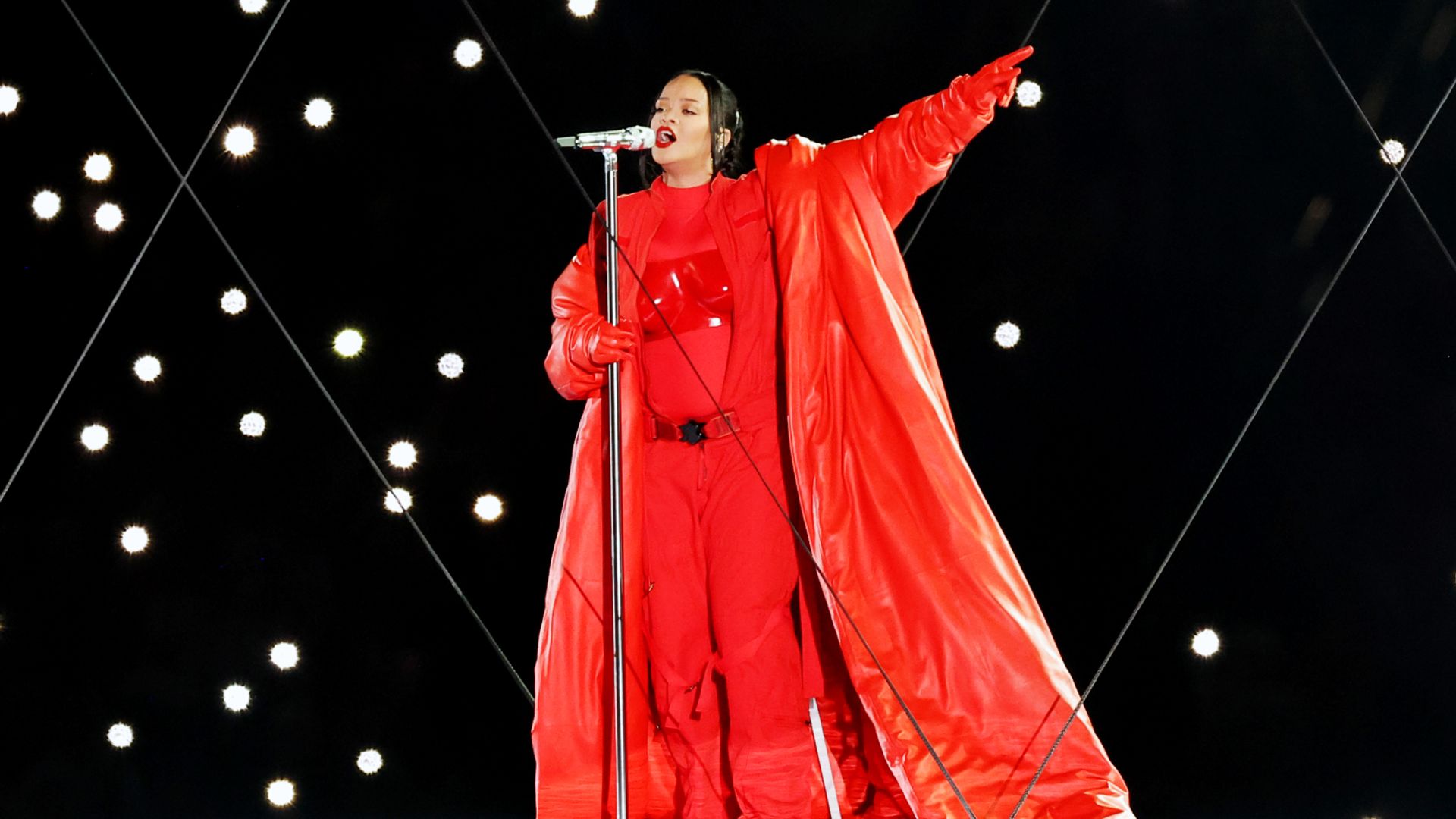Rihanna w jasnoczerwonej marynarce i długiej pelerynie występuje na scenie z mikrofonem