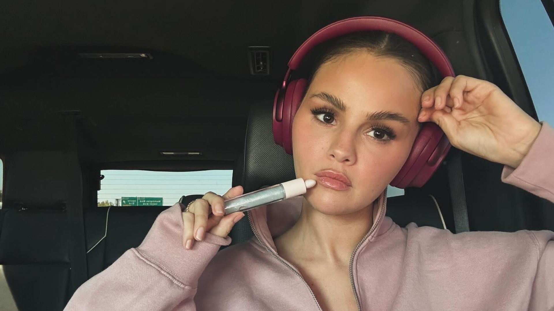 Selena Gomez w różowej bluzie i słuchawkach siedzi w samochodzie, trzymając przy twarzy produkt kosmetyczny