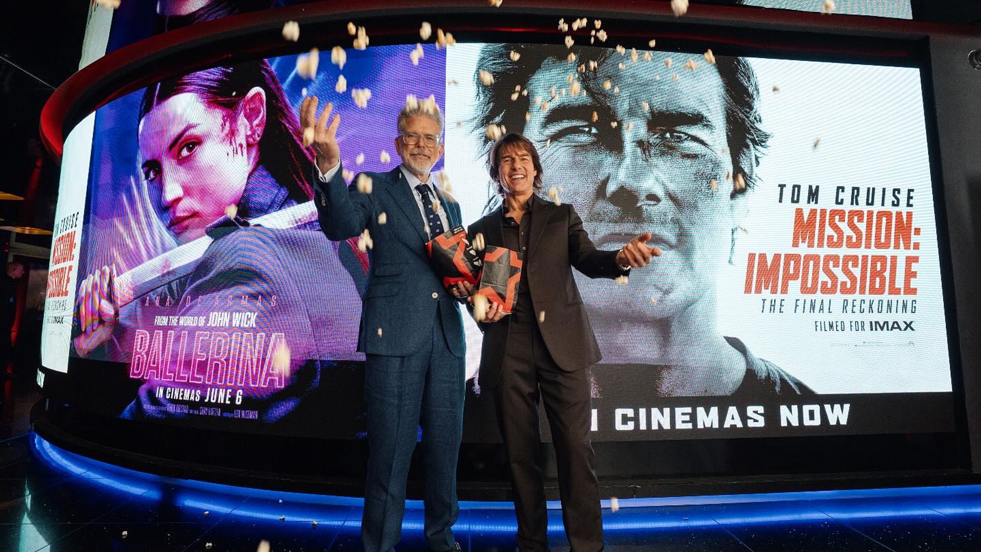 Dwóch mężczyzn w garniturach pozuje z pudełkami popcornu na tle plakatów filmowych Ballerina i Mission: Impossible