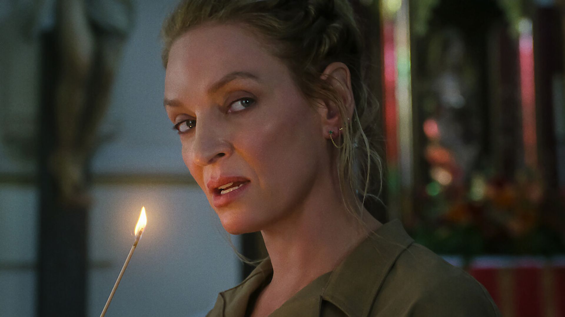 Uma Thurman z włosami zaczesanymi do tyłu trzyma zapaloną świecę i patrzy w bok na lekko rozmytym tle