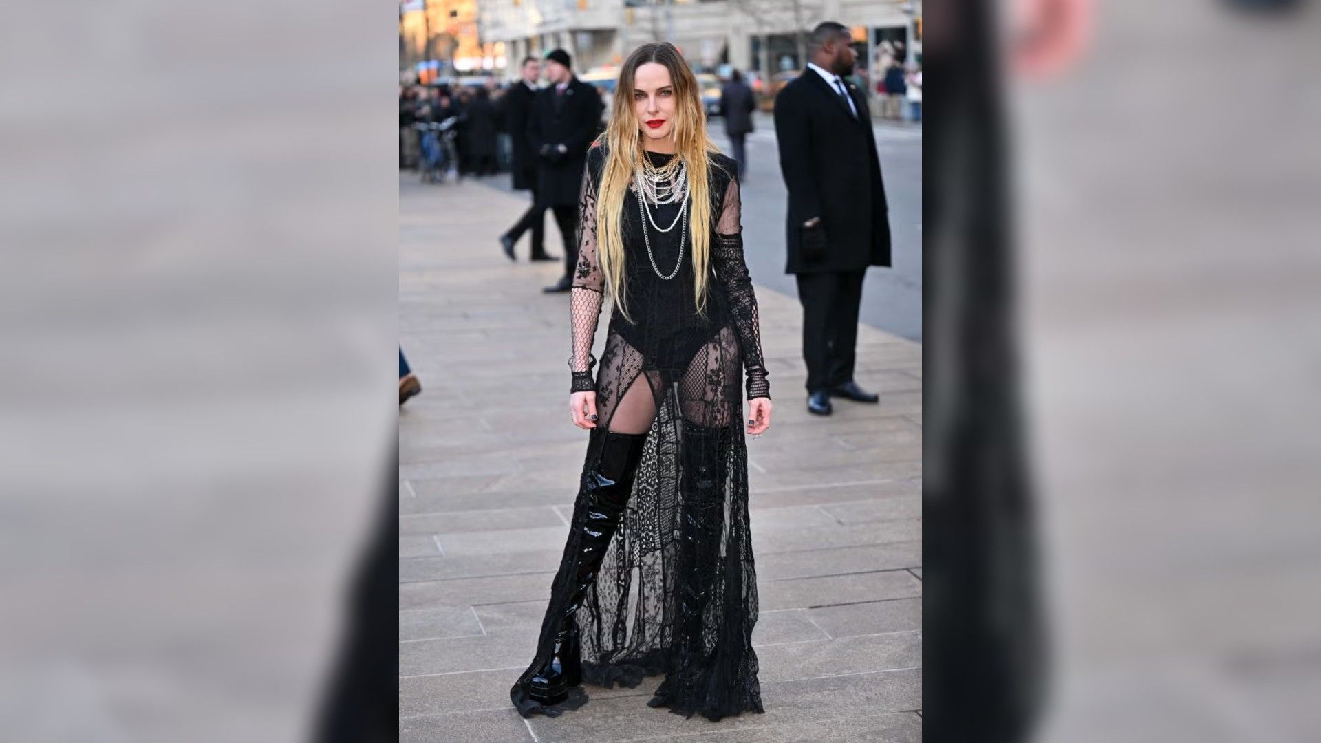 Rebecca Ferguson pozuje na zewnątrz w długiej, prześwitującej czarnej sukni z koronką i wysokich lakierowanych butach.