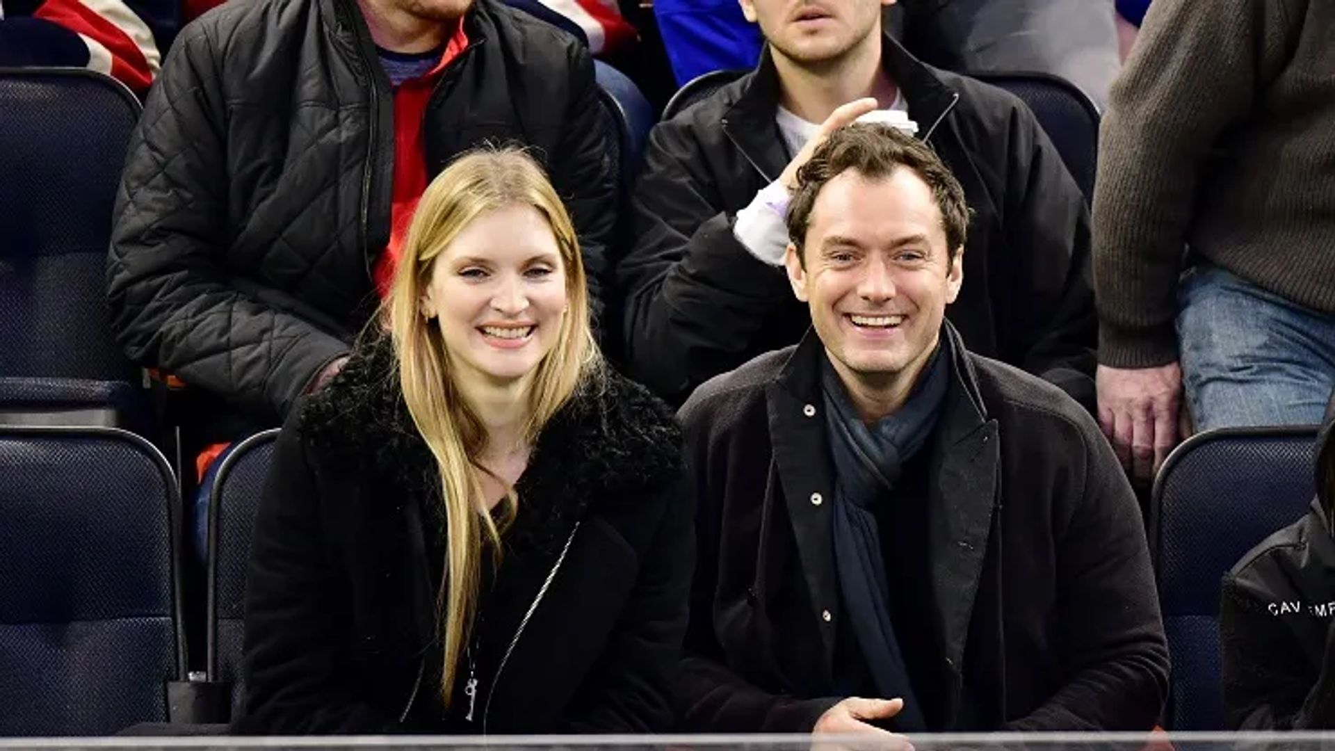 Jude Law i Phillipa Coan siedzą obok siebie na trybunach, uśmiechają się i patrzą w kamerę