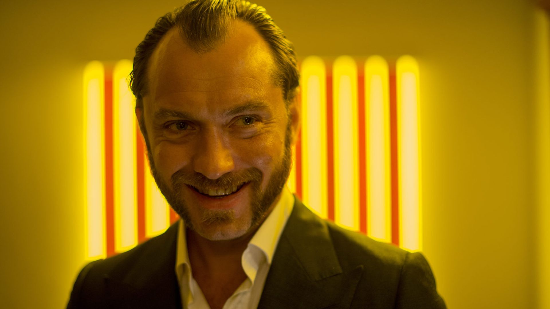 Jude Law w marynarce uśmiecha się na tle jasnych żółtych smug światła
