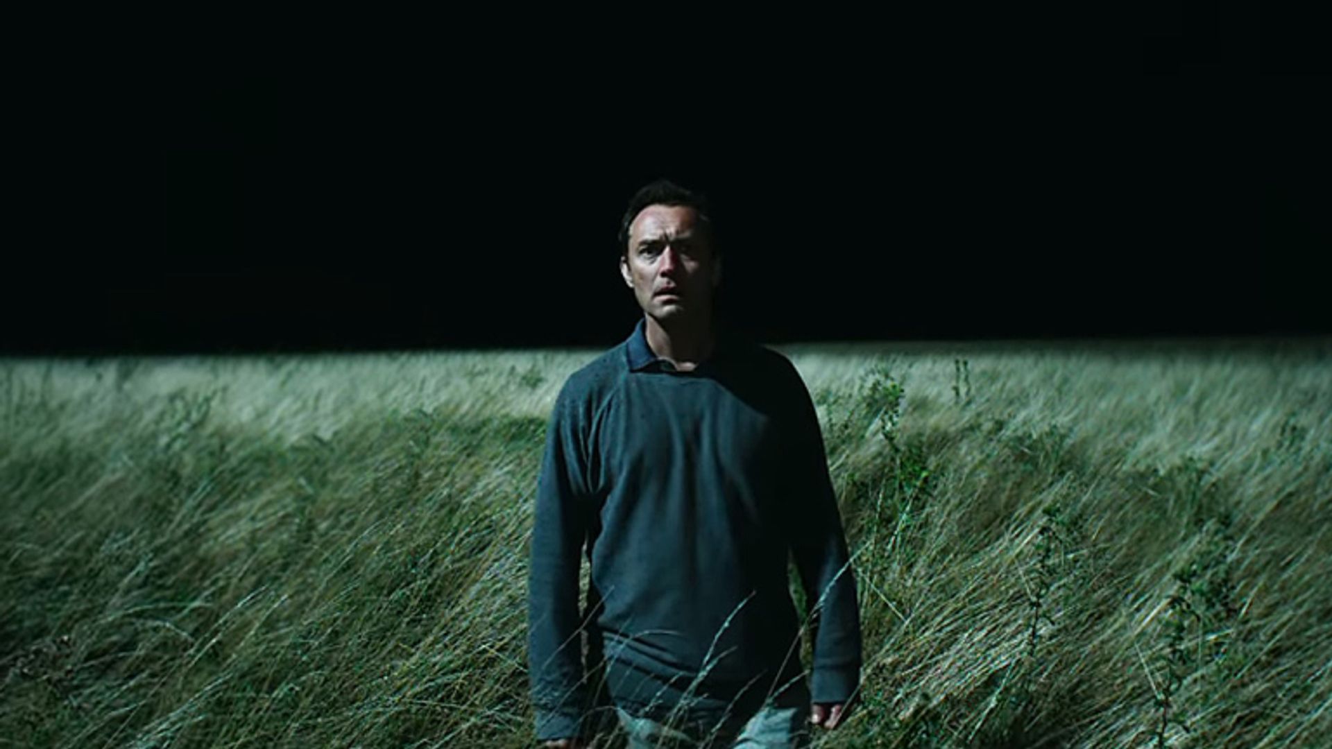 Jude Law w ciemnym ubraniu stoi w wysokiej trawie nocą, oświetlony ostrym światłem