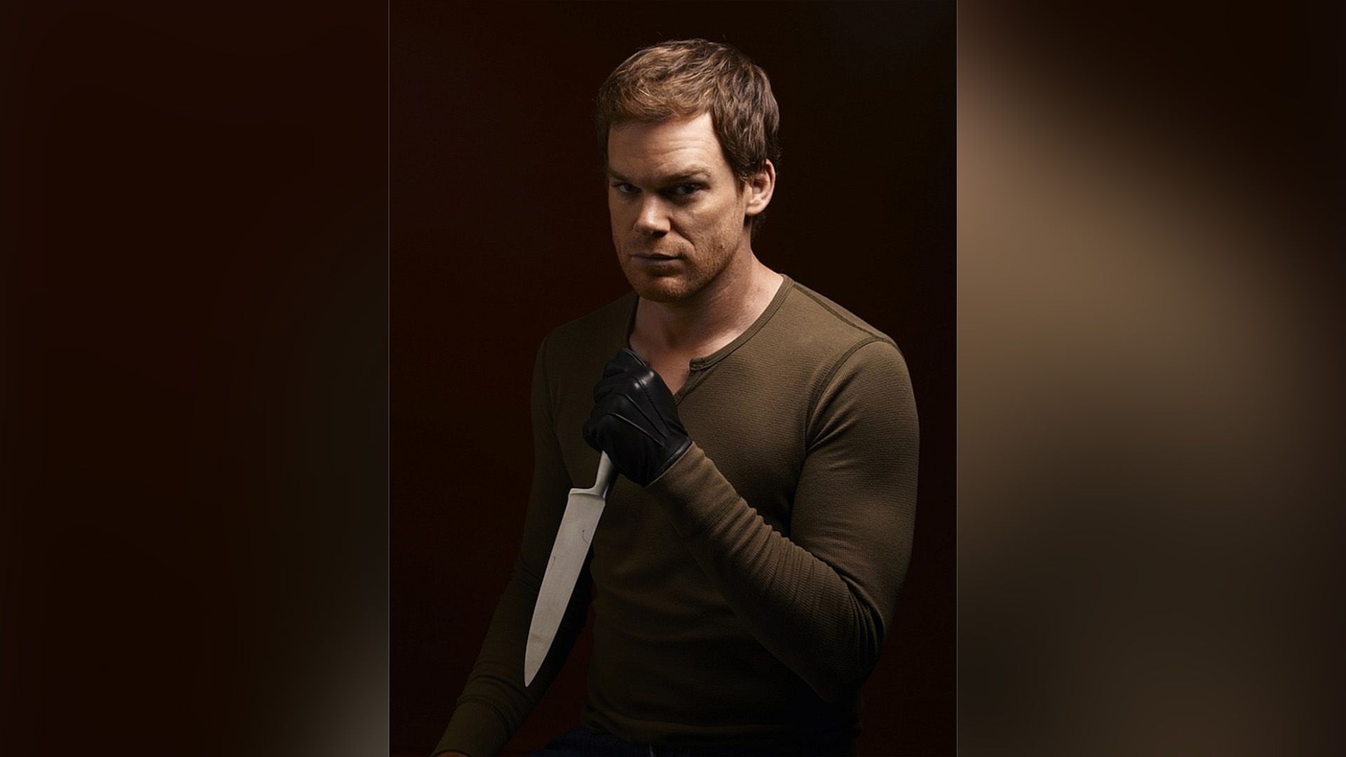 Michael C. Hall w brązowej kurtce i czarnych rękawiczkach trzymający nóż w dłoni
