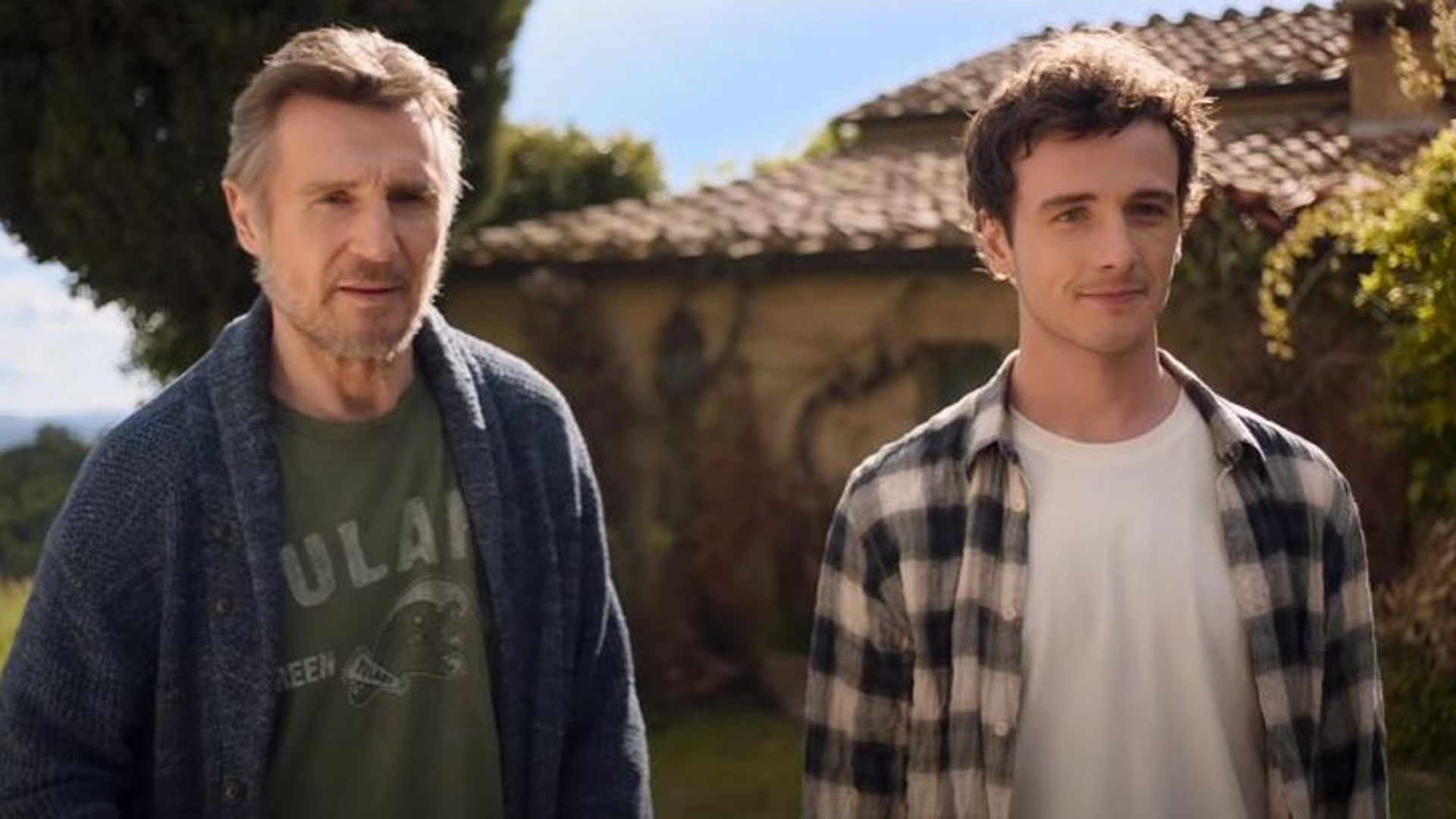 Liam Neeson i Michael Richardson stoją obok siebie przed domem z dachówkowym dachem w słoneczny dzień