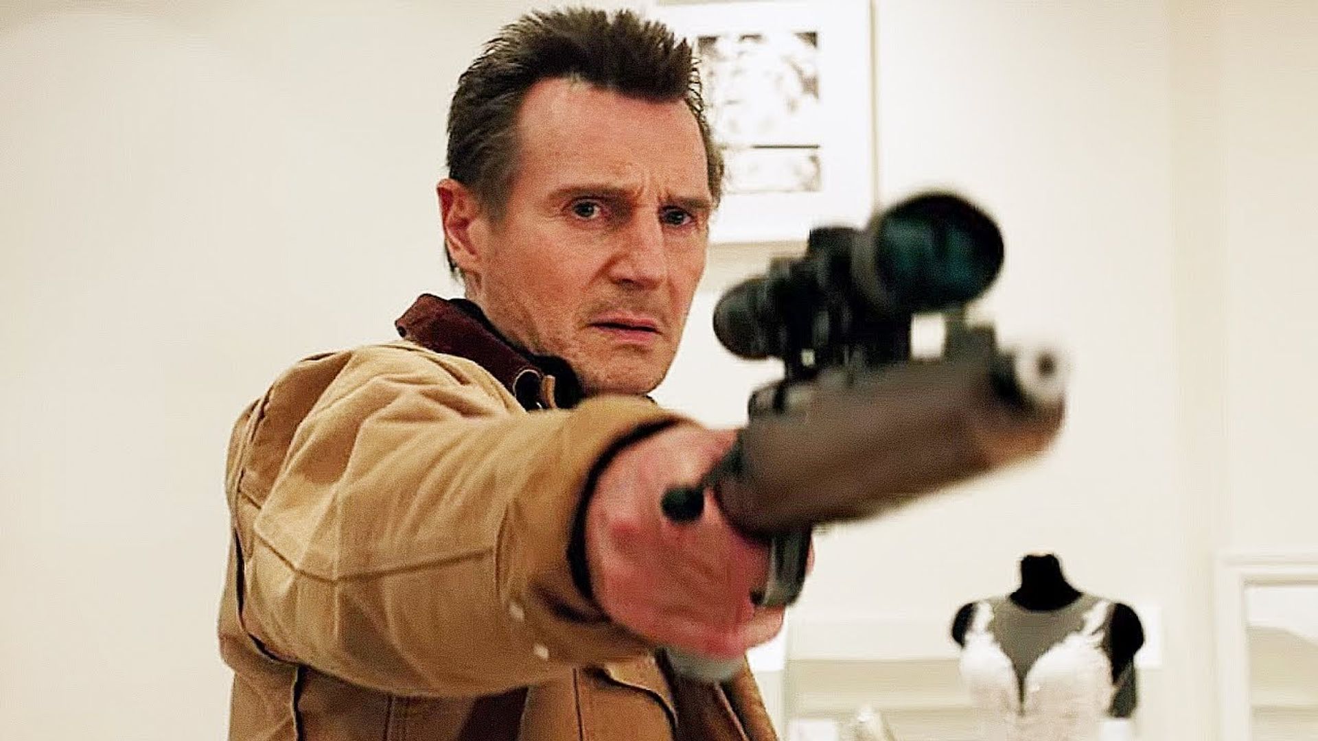 Liam Neeson w beżowej kurtce celuje z karabinu z lunetą, za nim manekin w sukni ślubnej