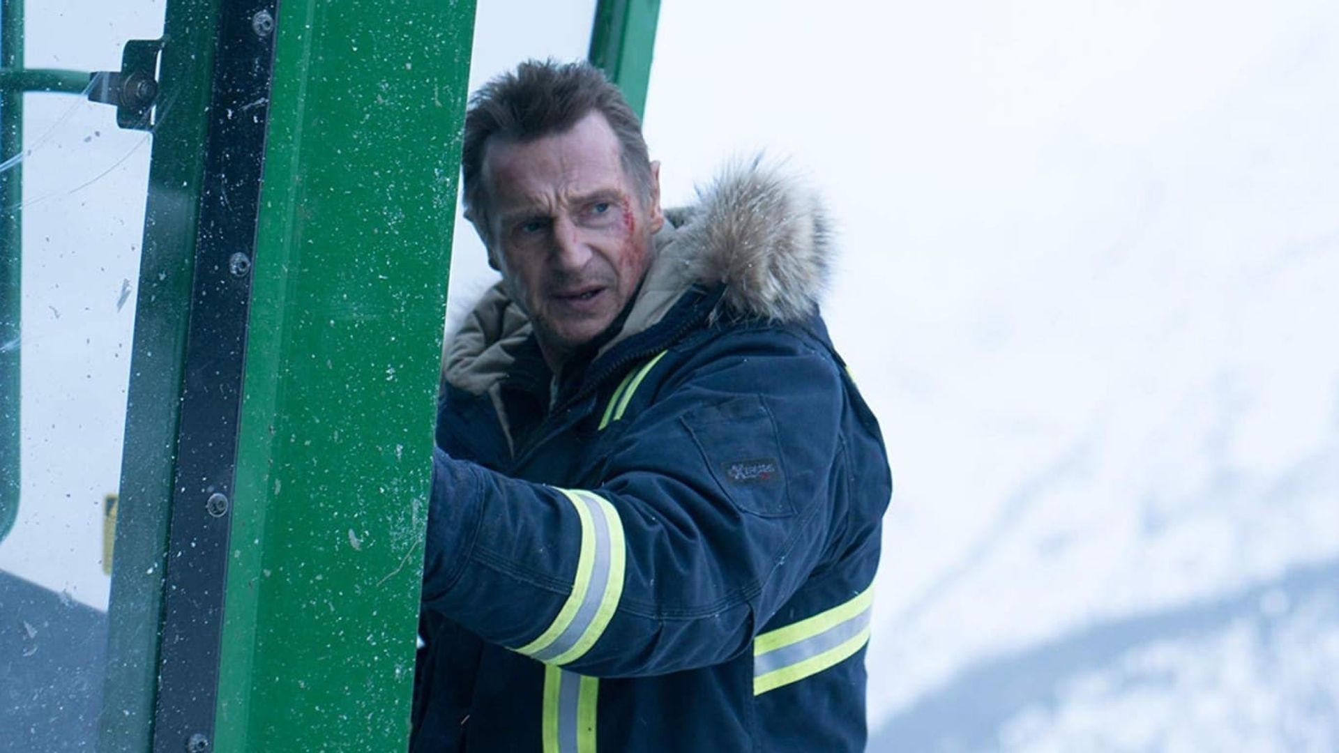 Liam Neeson w zimowej kurtce z kapturem trzyma drzwi zielonej kabiny na tle ośnieżonego krajobrazu