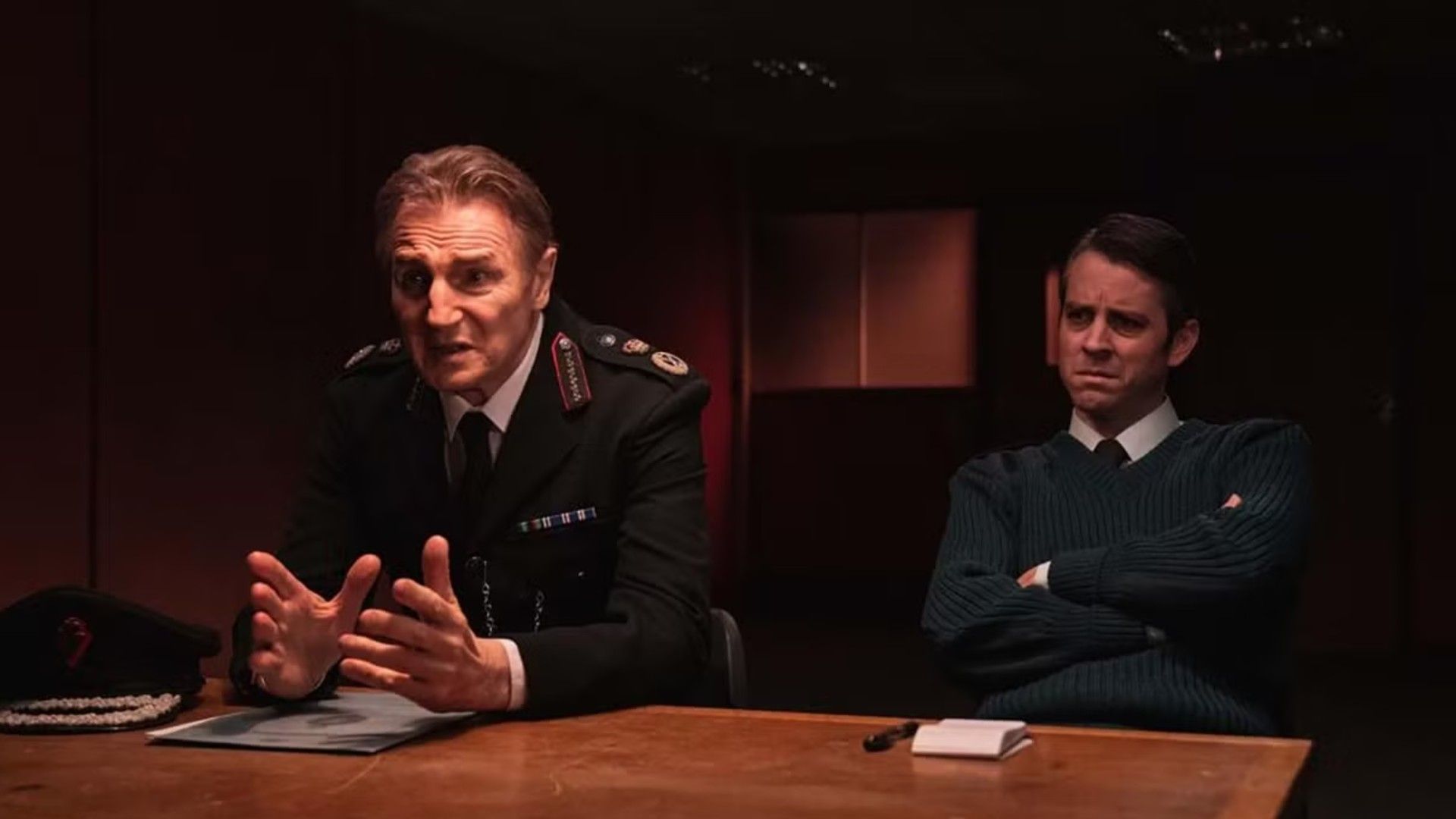 Liam Neeson w mundurze policyjnym siedzący przy stole przesłuchań, scena z serialu "Derry Girls"