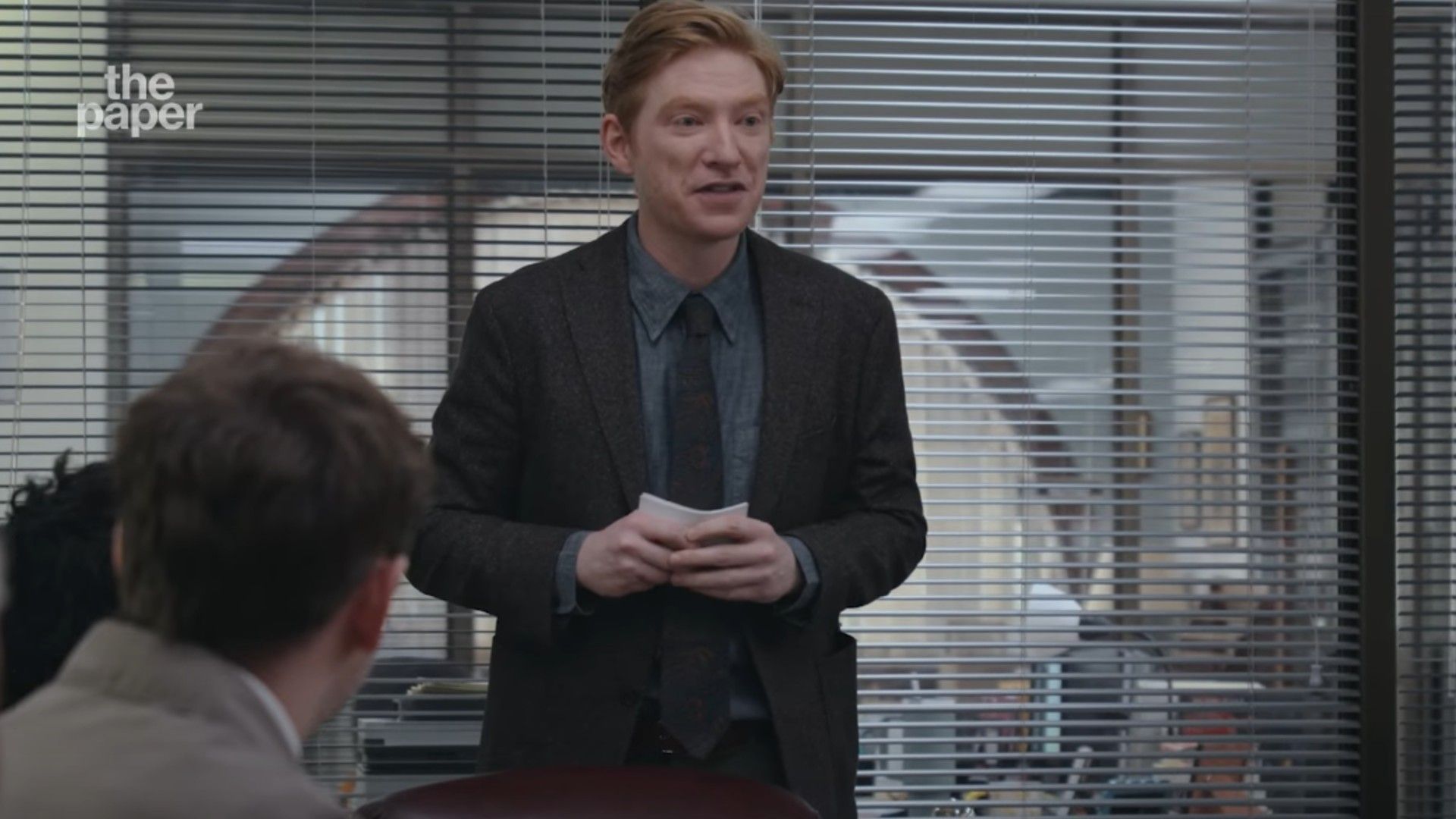 Domhnall Gleeson w garniturze stojący w sali konferencyjnej