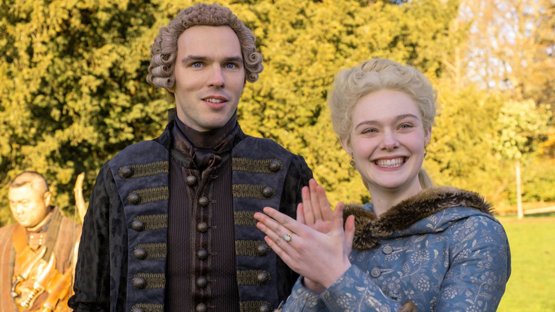 Nicholas Hoult i Elle Fanning w osiemnastowiecznych kostiumach stojący na ulicy, aktorka się uśmiecha i klaszcze w dłonie