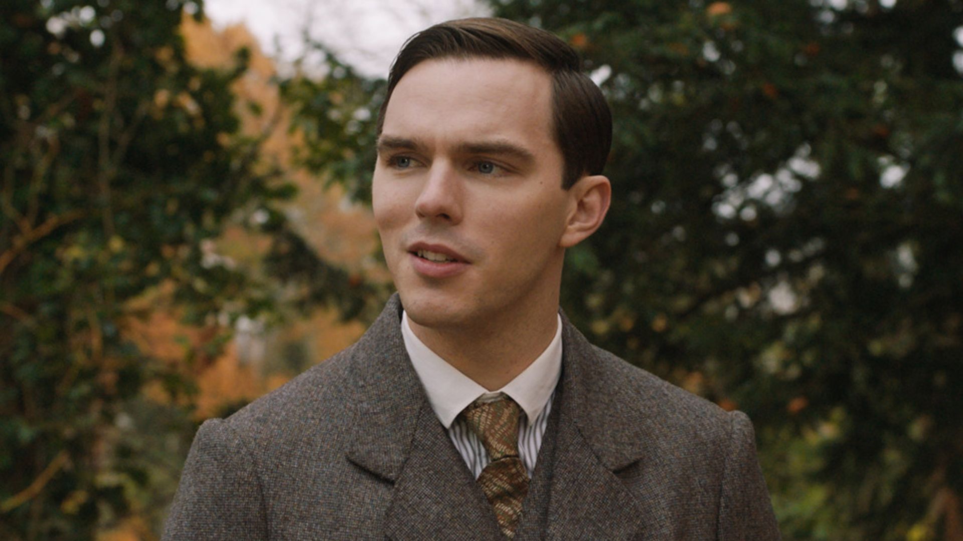 Nicholas Hoult w brązowym garniturze i krawacie stojący na ulicy w roli młodego J.R.R. Tolkiena