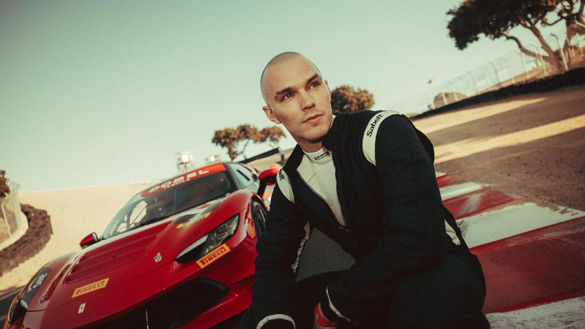 Nicholas Hoult w kombinezonie wyścigowym siedzi na torze z czerwonym sportowym Ferrari za sobą