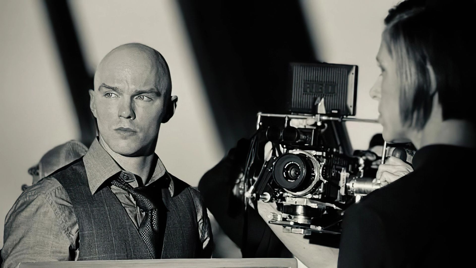 Nicholas Hoult w kamizelce i krawacie stoi obok profesjonalnej kamery filmowej na planie filmowym
