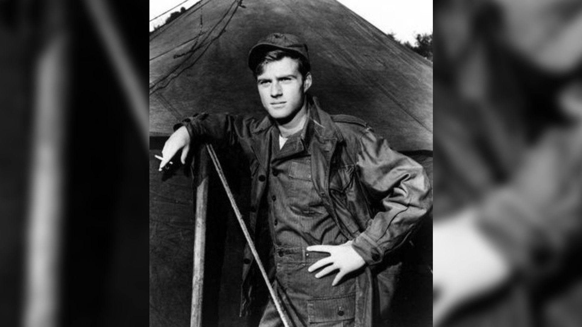 Czarno-biała fotografia młodego Roberta Redforda w mundurze wojskowym stojącego przy namiocie, scena z filmu "War Hunt"