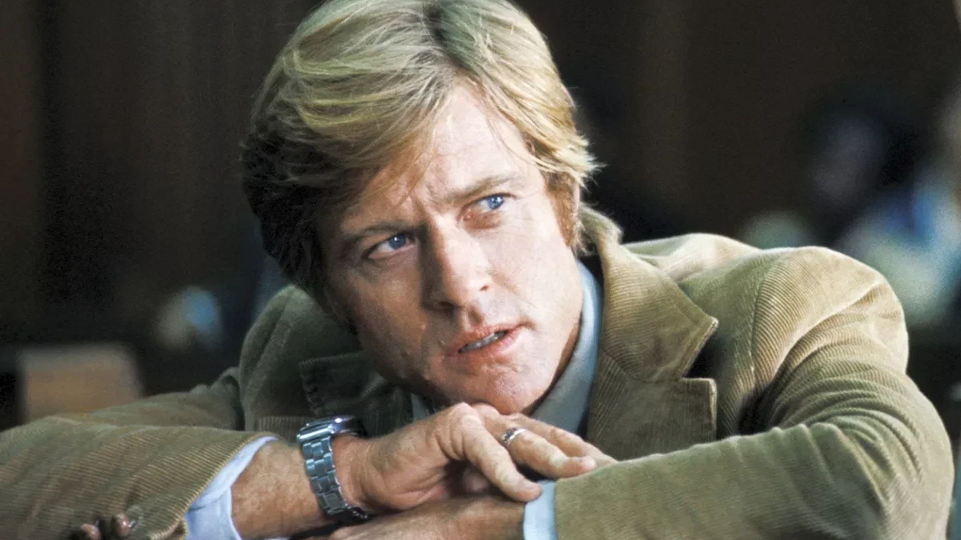 Robert Redford w brązowej marynarce z zegarkiem na nadgarstku, oparty na rękach i patrzący w bok
